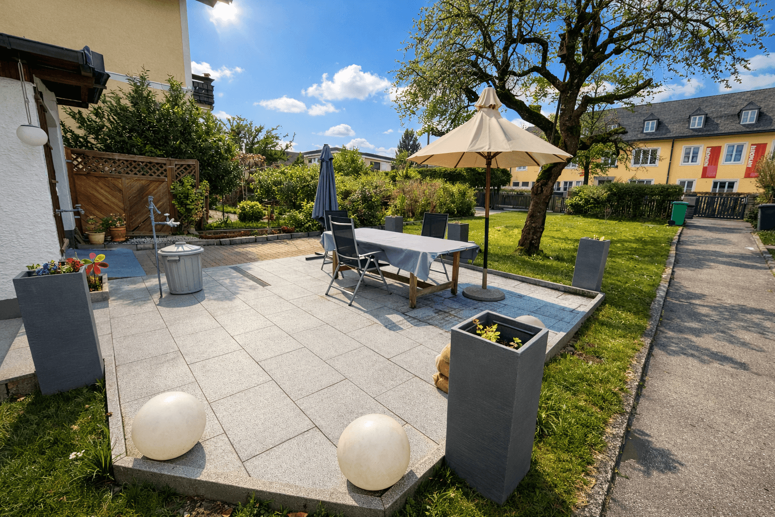 Sonniger Außenbereich mit gepflasterter Terrasse, Esstisch und Sonnenschirm im Garten.