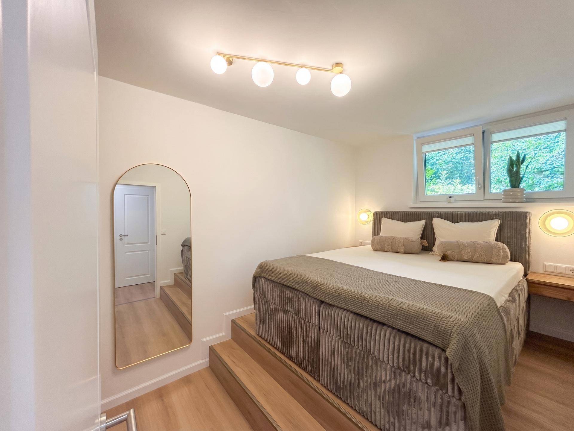 Helles Schlafzimmer mit erhöhtem Bett, Holzfußboden und großem Spiegel, bietet eine gemütliche Atmosphäre.