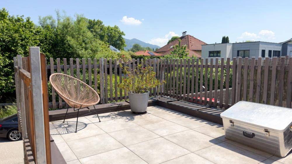Dachterrasse mit Fliesenboden, Sitzgelegenheit und Holzzaun unter blauem Himmel.