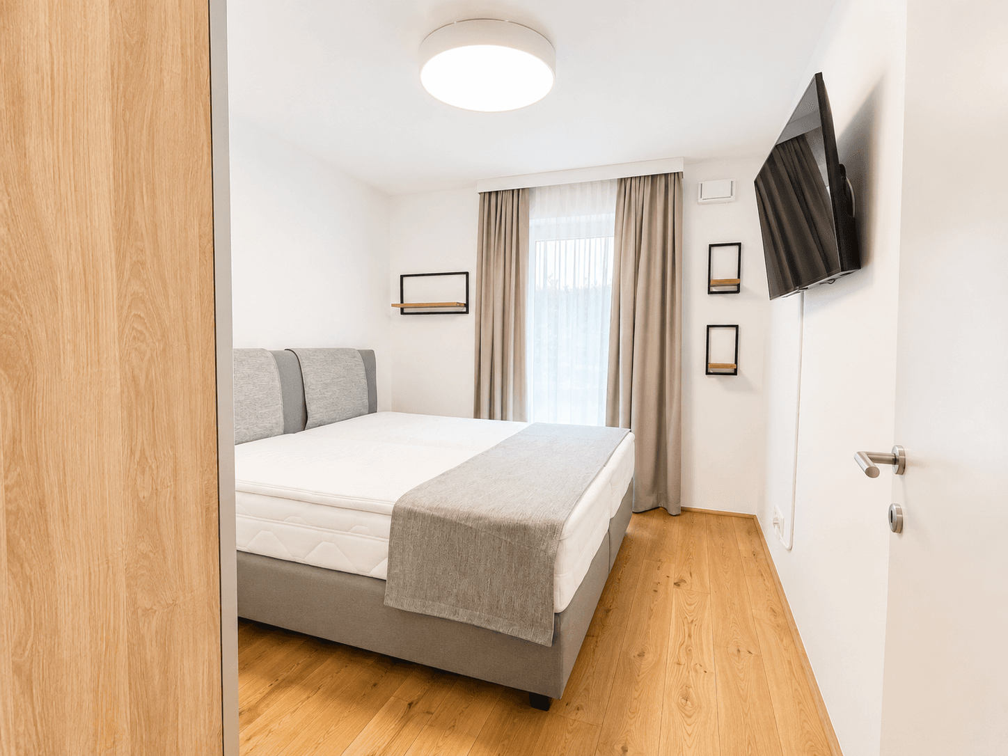 Helles Schlafzimmer mit Doppelbett, Wandfernseher und modernem Holzboden für angenehmen Wohnkomfort.