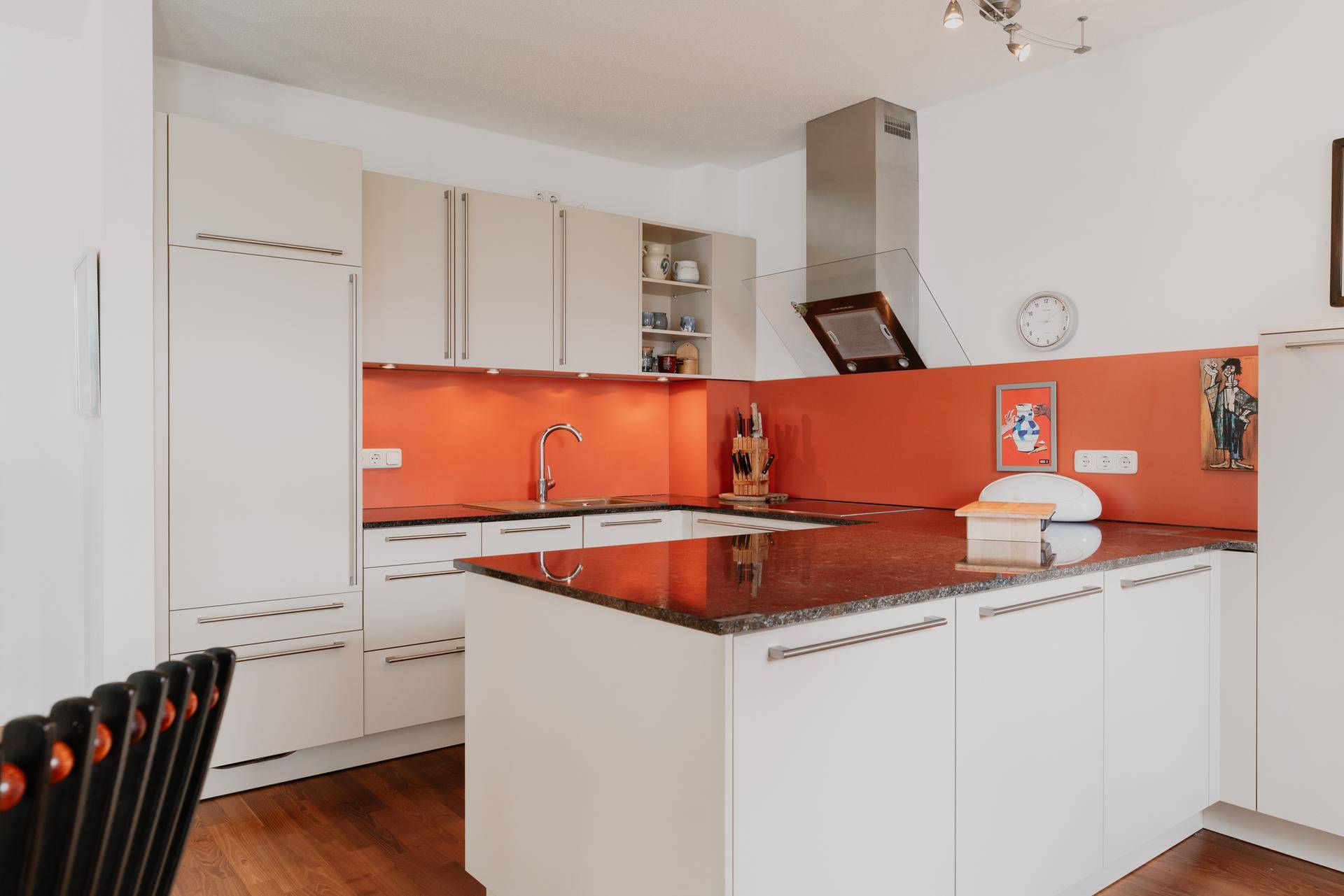Moderne Einbauküche mit weißen Fronten, Granitarbeitsplatten und orangefarbener Rückwand.