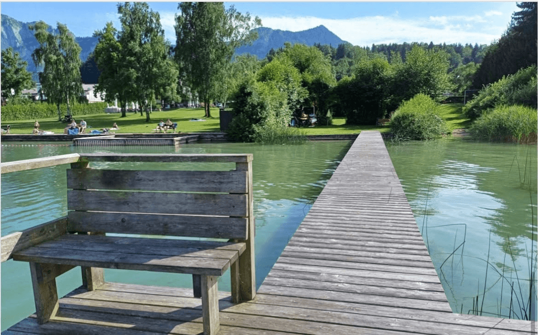 Idyllischer Blick auf einen See mit Holzsteg und Sitzbank, umgeben von grünen Bäumen und Bergen.