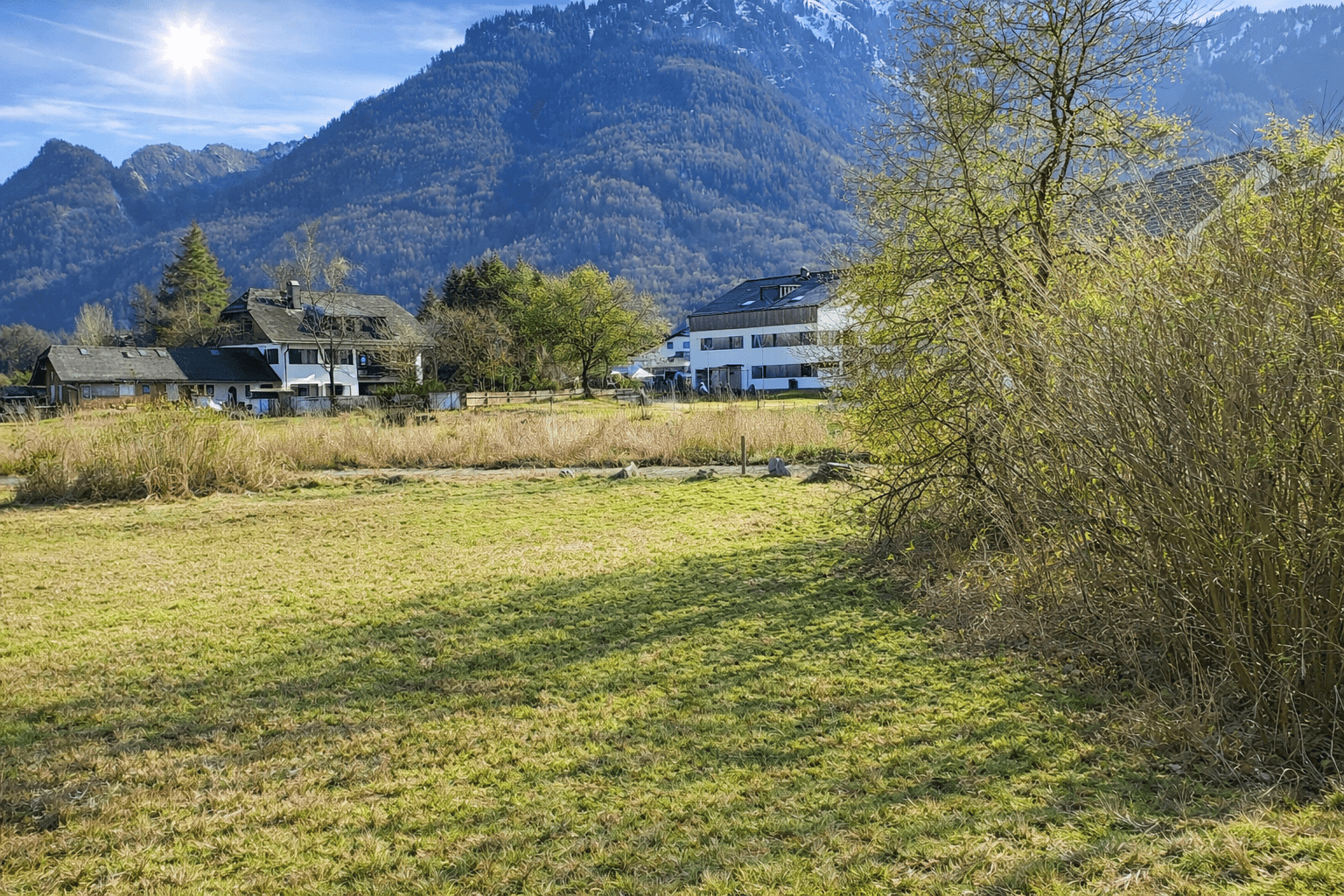 Sonnige Wiese mit Blick auf mehrere Wohnhäuser und die majestätische Bergkette im Hintergrund.