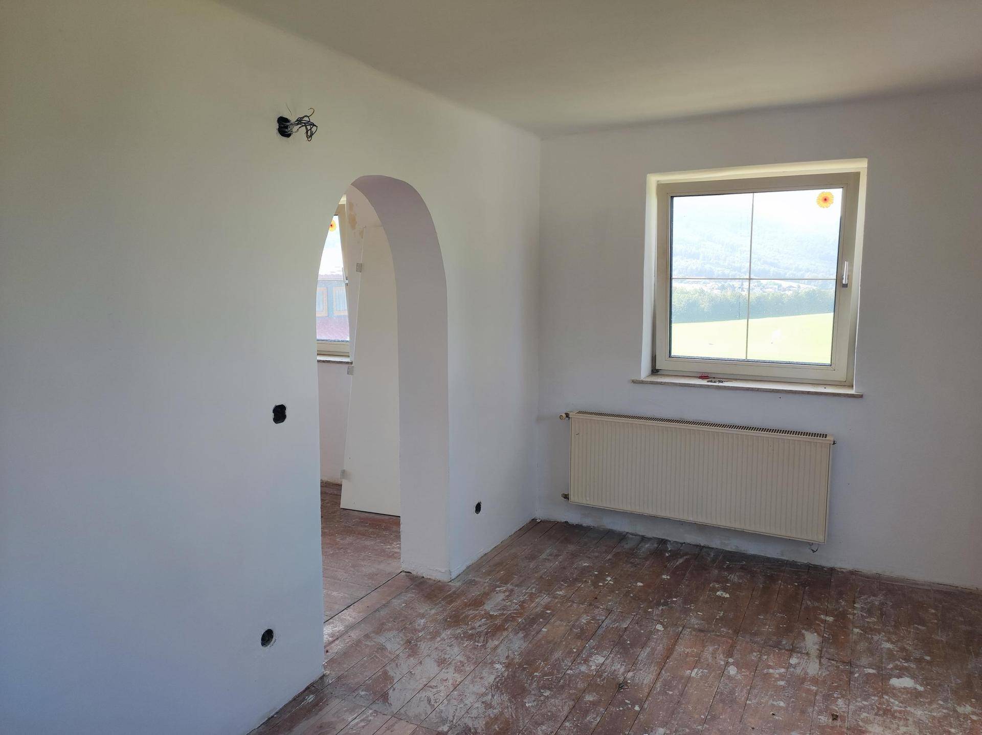 Unmöblierter Raum mit Fenster, Heizkörper und abgenutztem Holzboden, der auf Renovierung wartet.