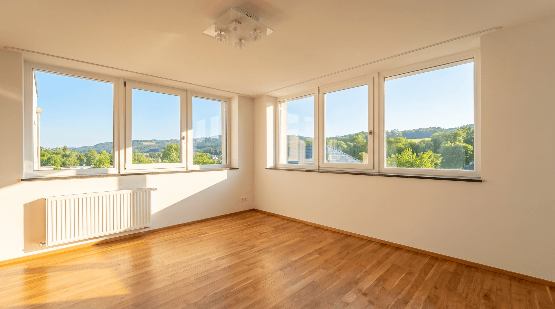 Helles, leeres Zimmer mit Holzboden und großen Fenstern, die einen weiten Blick ins Grüne bieten.