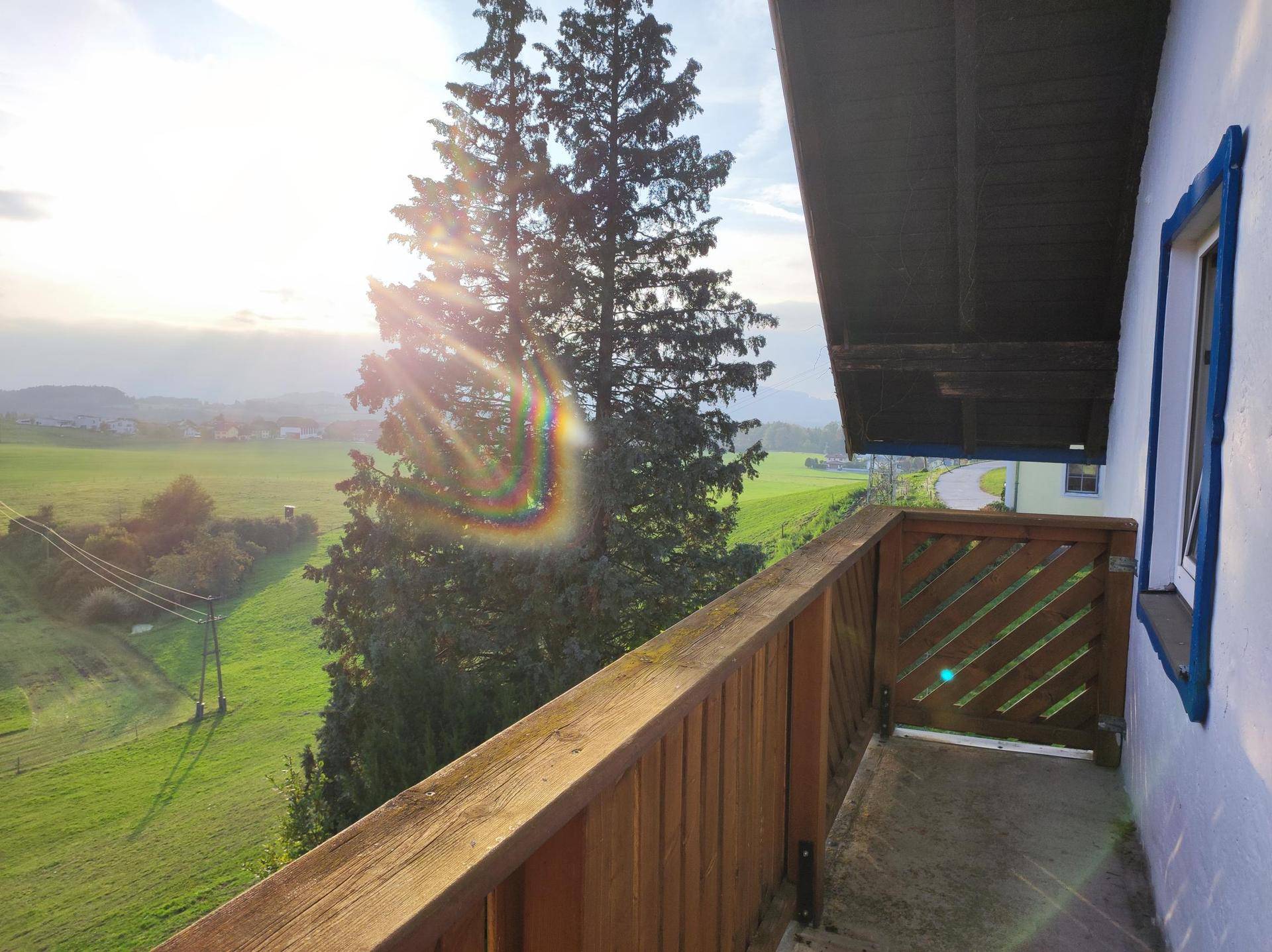 Sonniger Balkon mit Holzgeländer und weitem Blick über grüne Felder und die umliegende Landschaft.