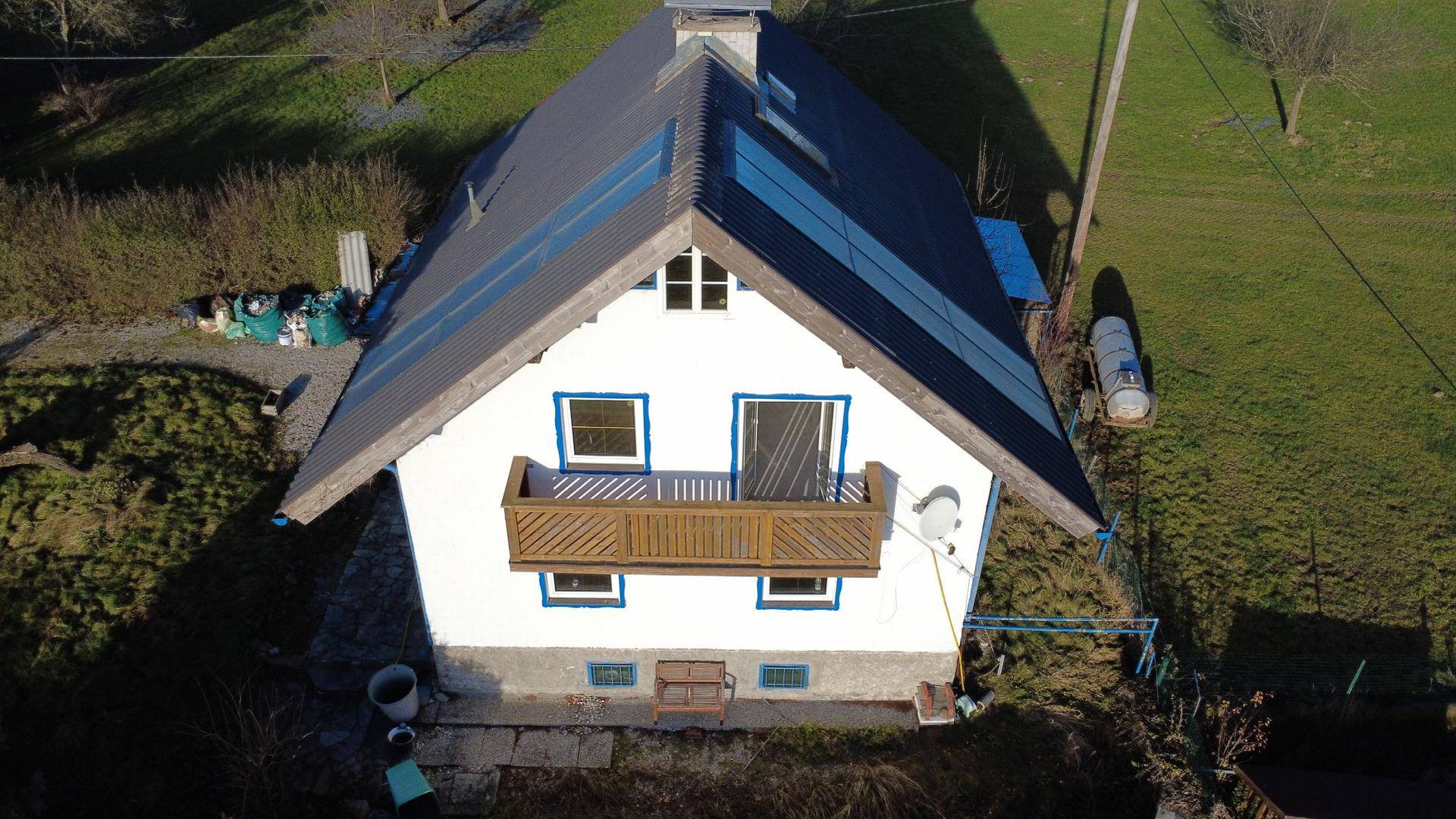 Luftaufnahme eines Hauses mit Solarmodulen auf dem Dach und einem Holzbalkon, eingebettet in grüne Landschaft.