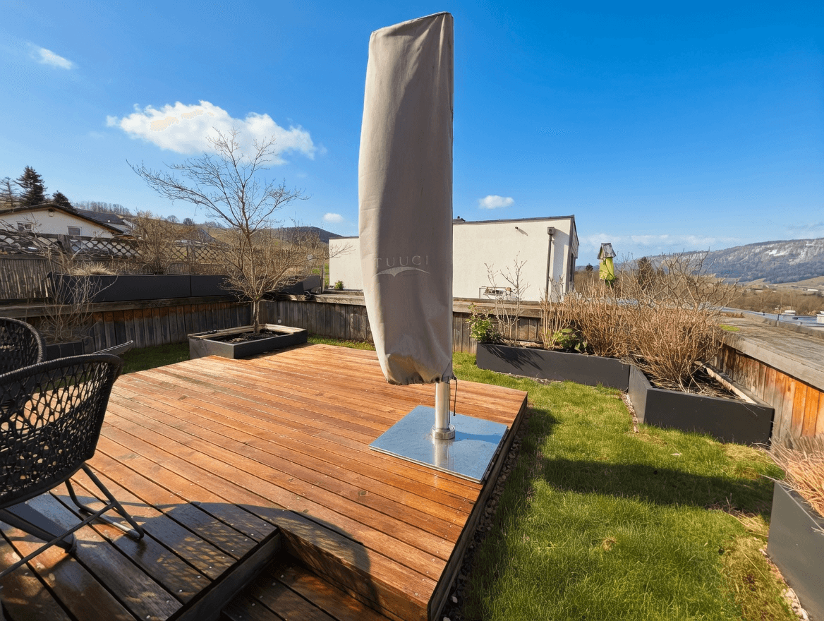 Sonniger Terrassenbereich mit Holzdeck, Rasenfläche und Pflanzen in Hochbeeten, ideal zum Entspannen.