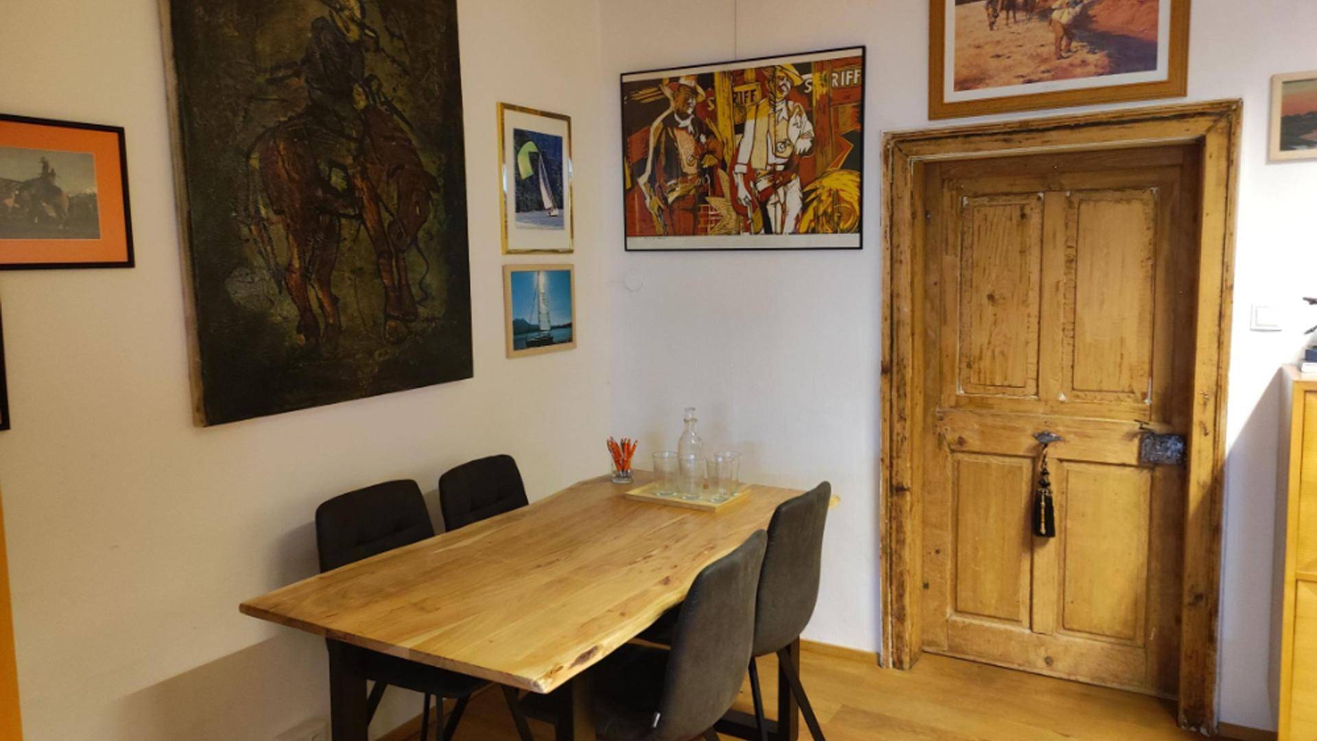 Essbereich mit einem Holztisch, vier Stühlen und Kunstwerken an den Wänden, neben einer alten Holztür.