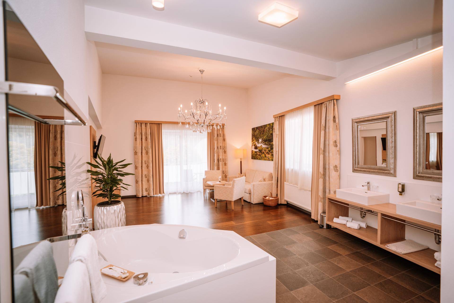 Geräumige Suite mit freistehender Badewanne, Doppelwaschtisch und elegantem Wohnbereich unter einem Kronleuchter.