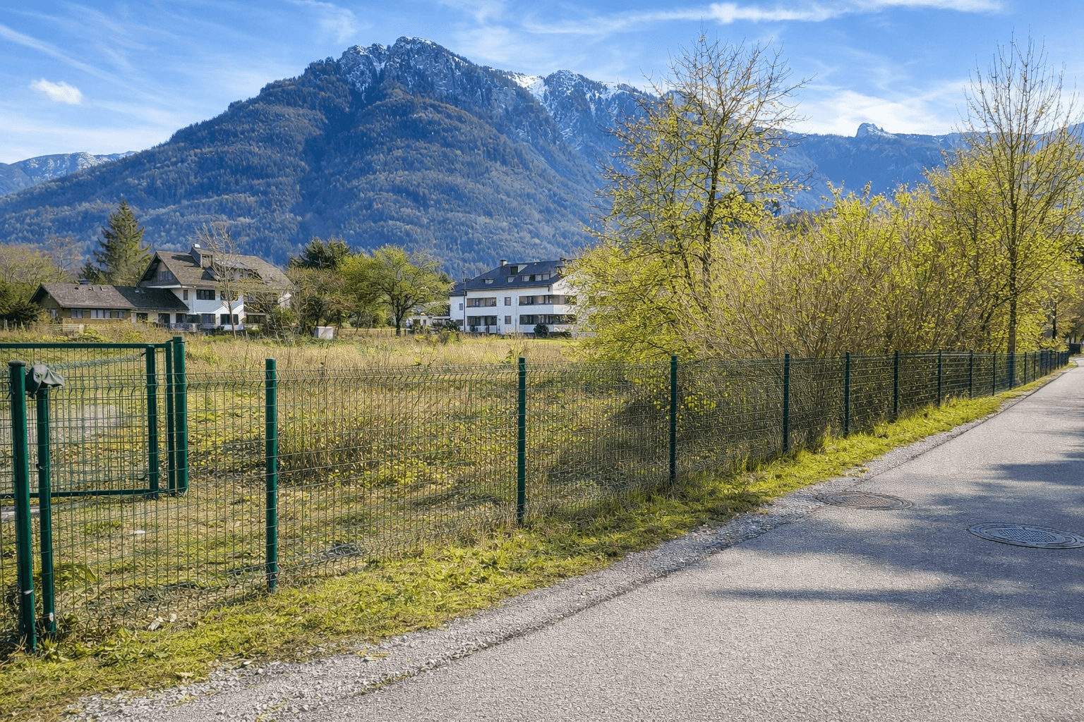 Grundstück mit Zaun und Straße, umgeben von grüner Natur und Bergen im Hintergrund.