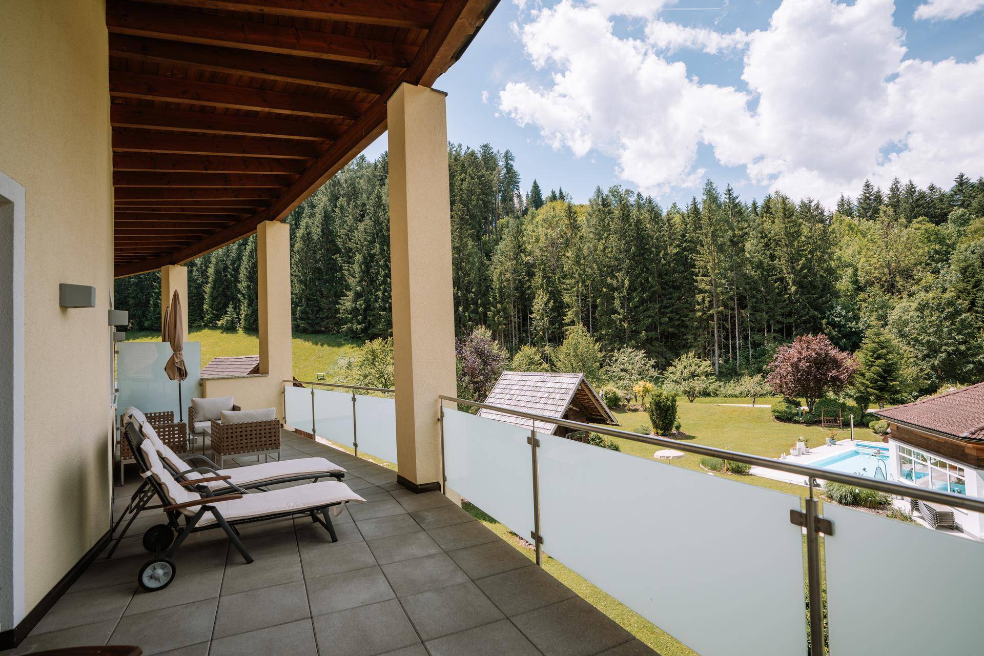 Sonniger Balkon mit Liegestühlen und Panoramablick auf den Wald, die Berge und den Poolbereich.