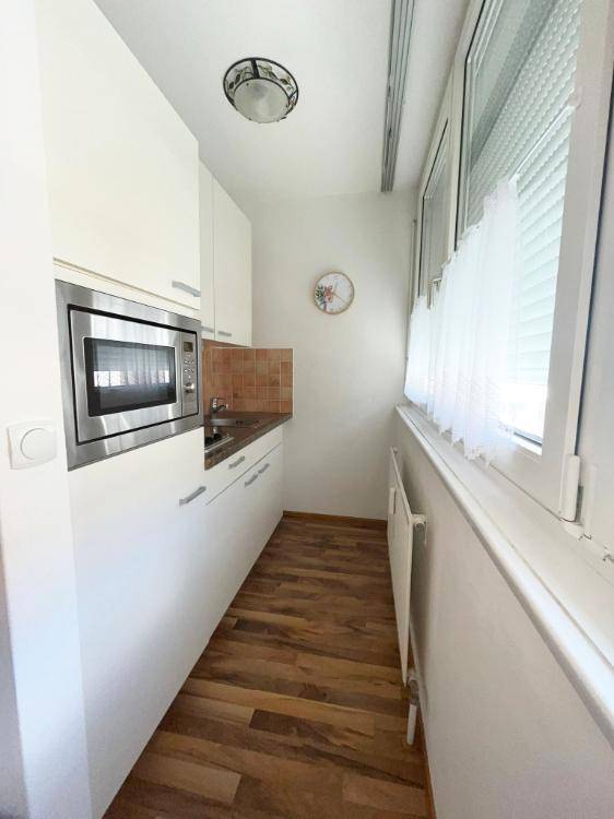 Schmale Küche mit weißen Schränken, integrierter Mikrowelle und Fenster für Tageslicht beim Kochen.