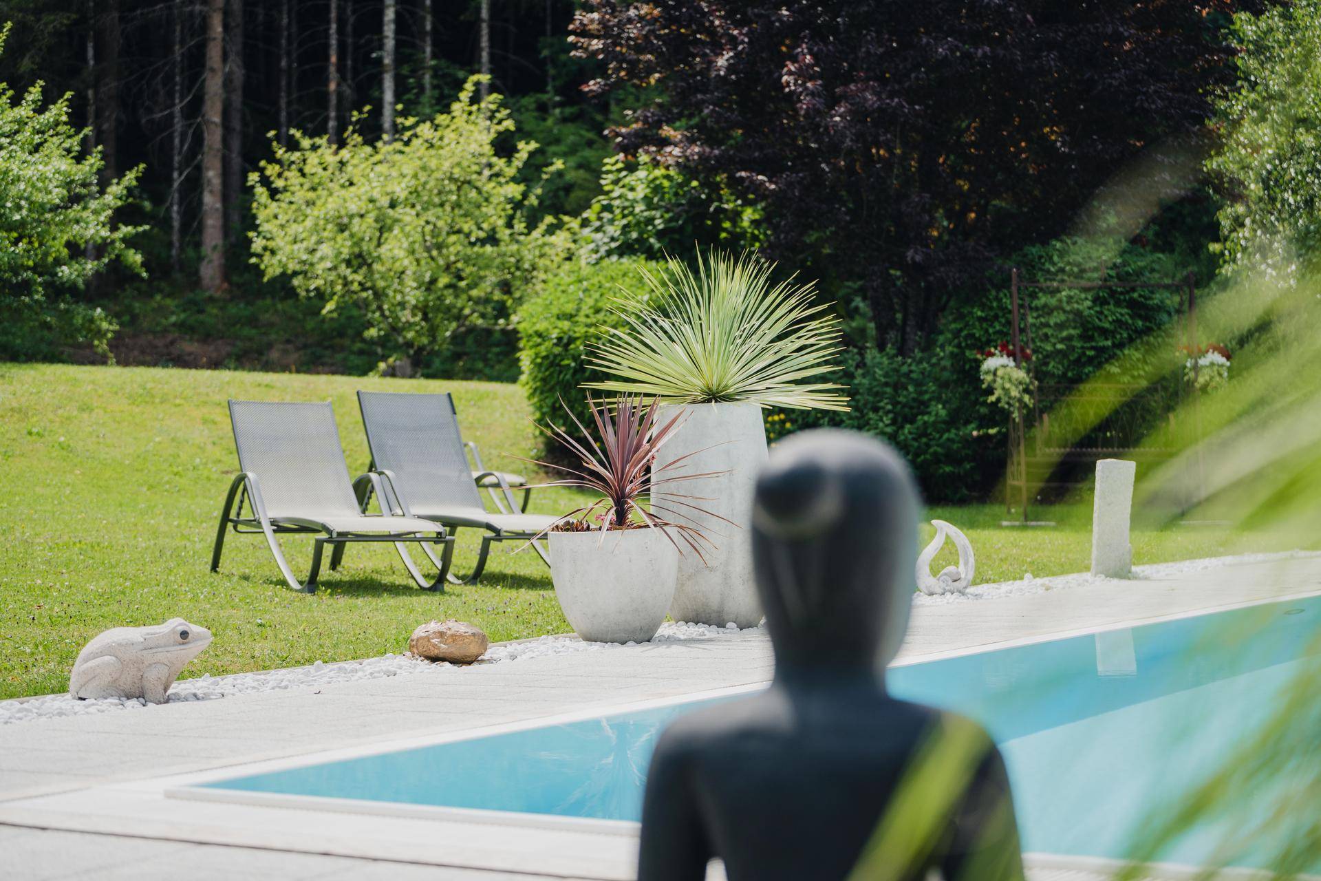 Entspannender Außenpoolbereich mit Liegestühlen, dekorativen Pflanzen und einer Statue im Grünen.