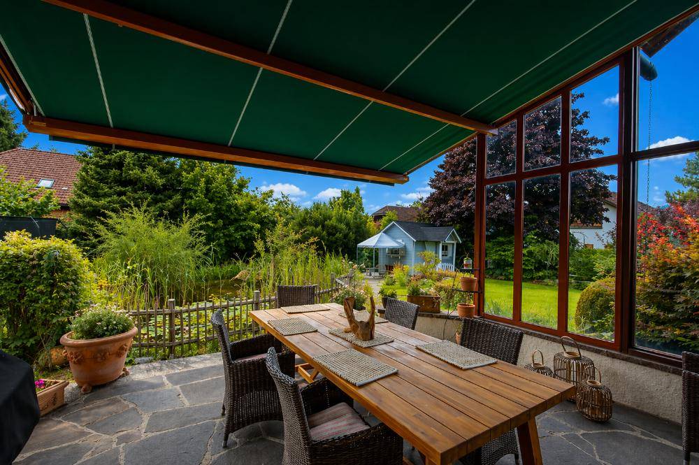 Überdachter Essbereich im Freien mit Holztisch und Blick auf den Garten mit Teich.