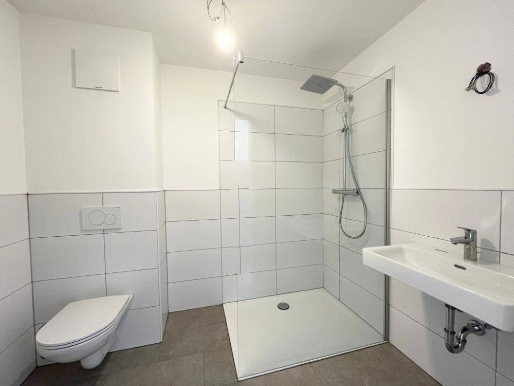 Funktionales Badezimmer mit moderner Dusche, Hänge-WC und Waschbecken in weiß gefliestem Raum.