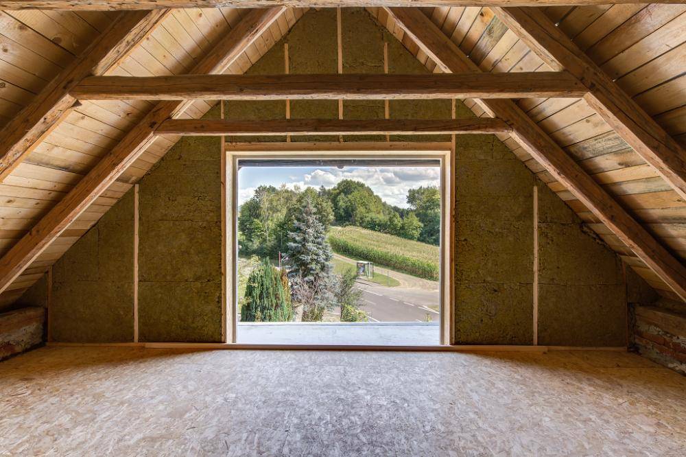 Ausbaufähiger Dachboden mit sichtbaren Holzbalken und großem Fenster mit Landschaftsblick.