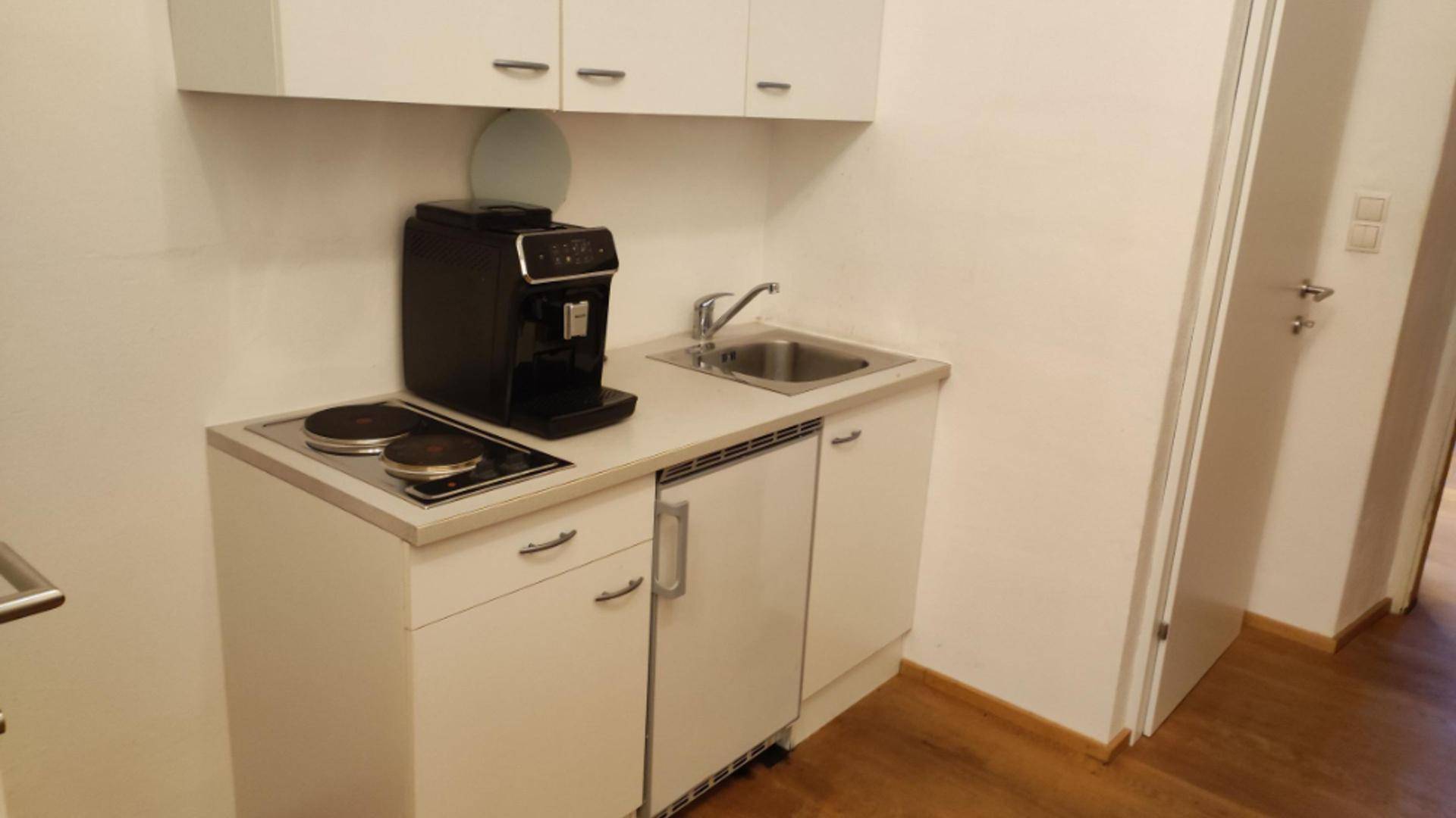Kleine Küchenzeile mit Spüle, zwei Kochplatten, einer Kaffeemaschine und weißen Schränken.