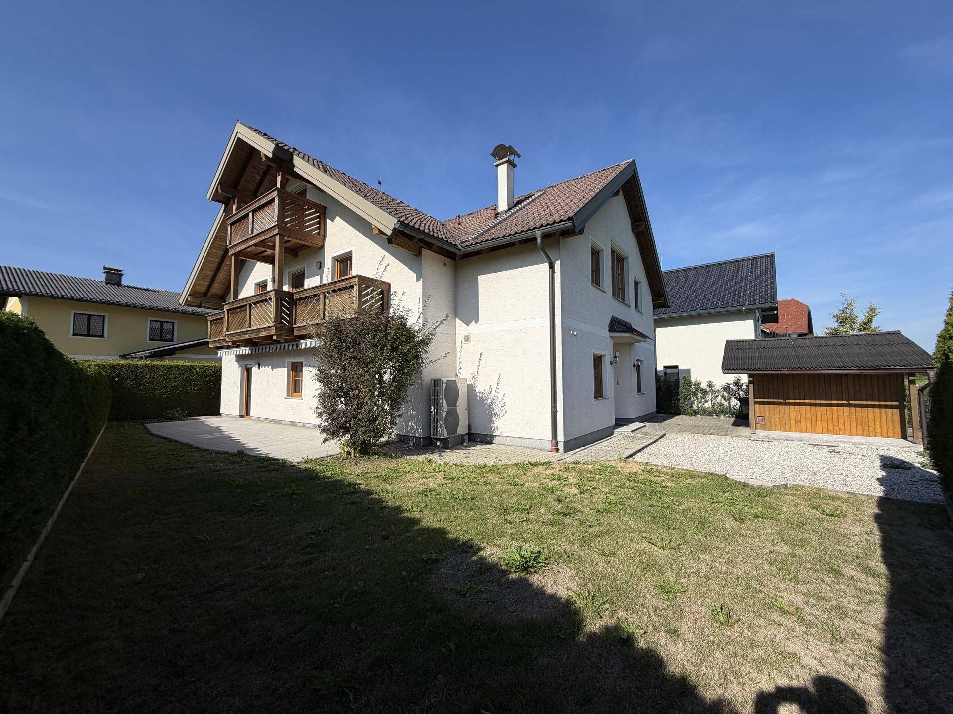 Gepflegtes Einfamilienhaus mit Garten, Terrasse, Holzbalkonen und separatem Carport unter blauem Himmel.