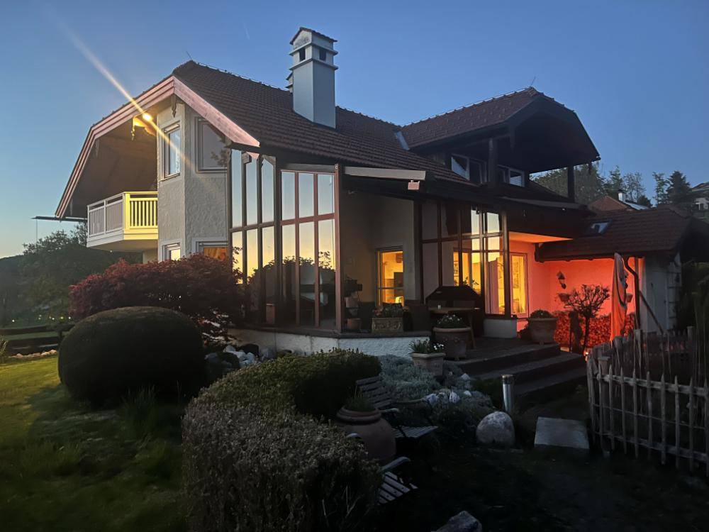 Hausfassade bei Sonnenuntergang mit beleuchteten Innenräumen und großen Glasflächen im Gartenbereich.