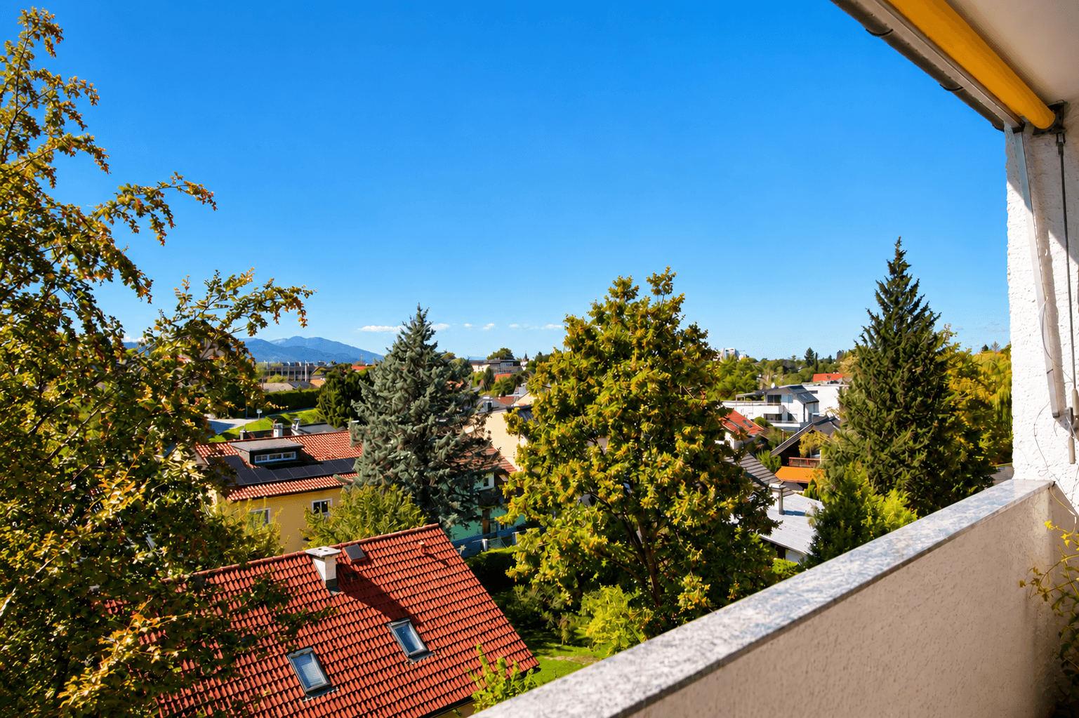 Weitläufiger Ausblick vom Balkon auf die grüne Umgebung, Dächer und ferne Berge.