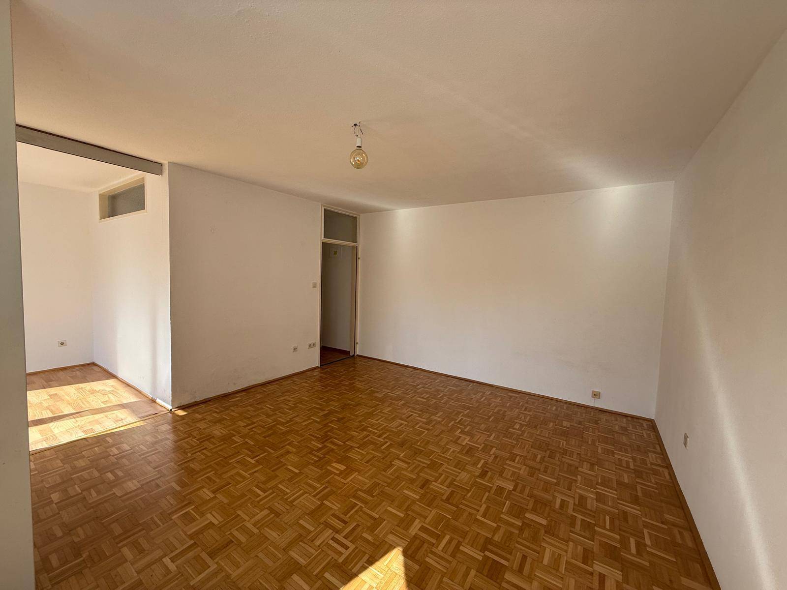 Geräumiges, leeres Zimmer mit Fischgrätparkett und viel natürlichem Licht.