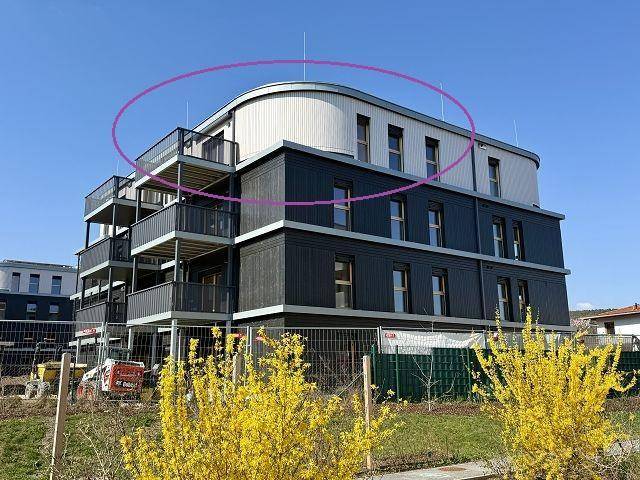 Modernes Mehrfamilienhaus mit dunkler Fassade und geschwungenem Dach, umgeben von Baustellenaktivitäten.