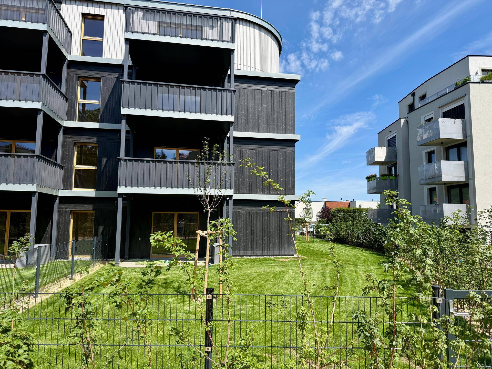Moderne Wohngebäude mit dunkler Holzfassade, Balkonen und gepflegter Grünfläche unter blauem Himmel.
