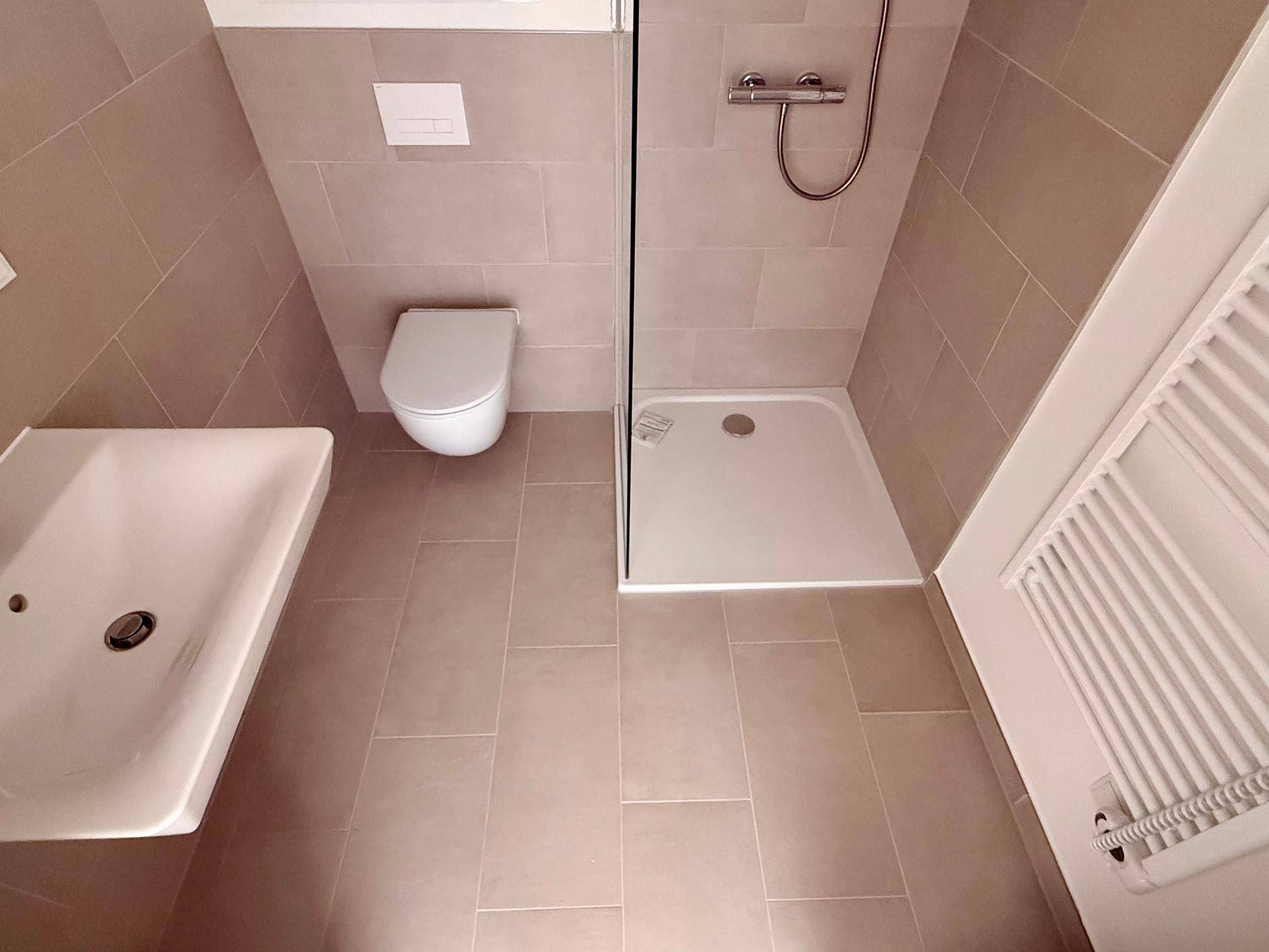 Badezimmer mit beigefarbenen Bodenfliesen, Duschbereich und wandhängendem WC.