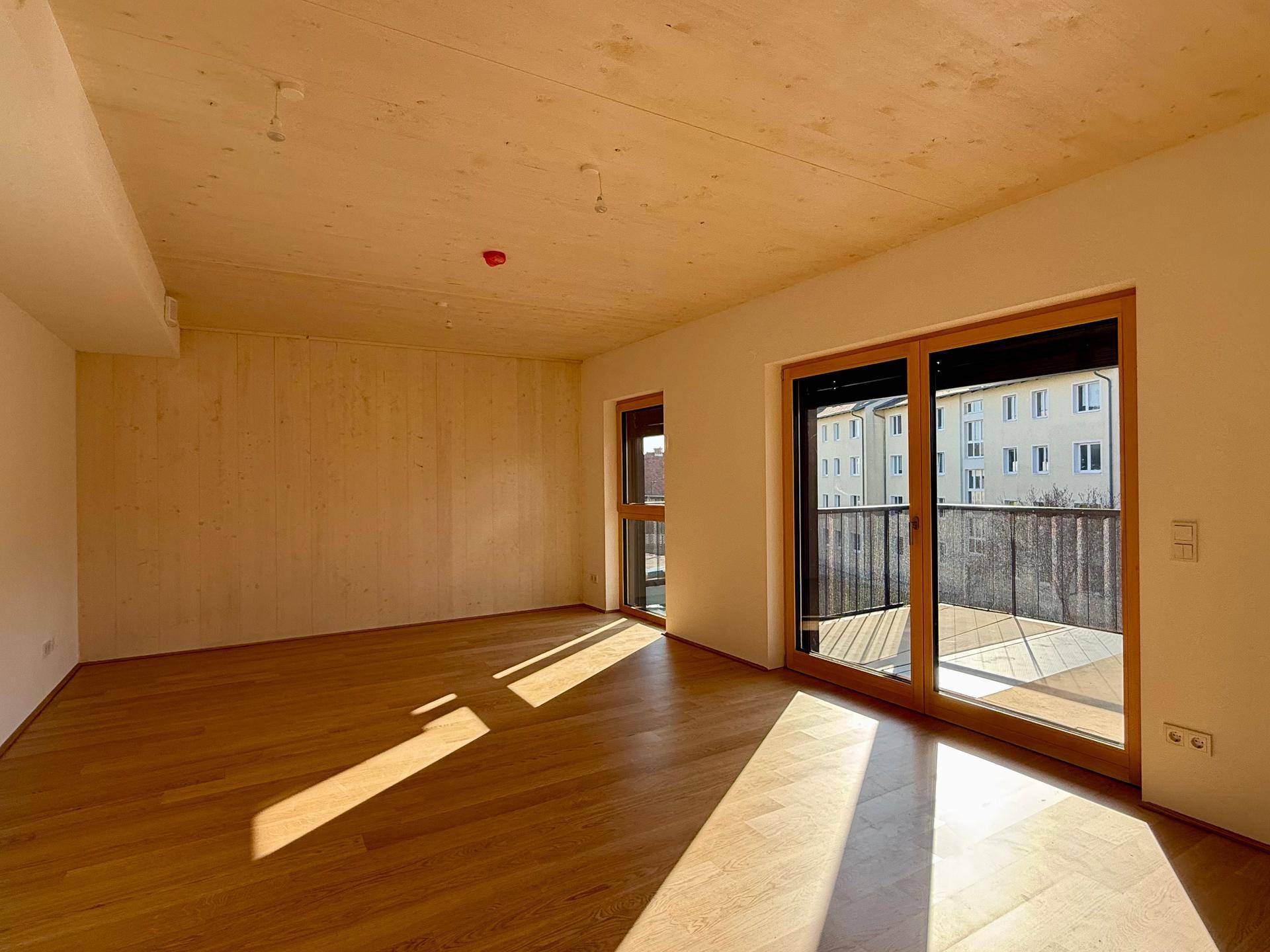 Helles Wohnzimmer mit Holzboden und Holzwand, großer Fensterfront und Zugang zum Balkon.