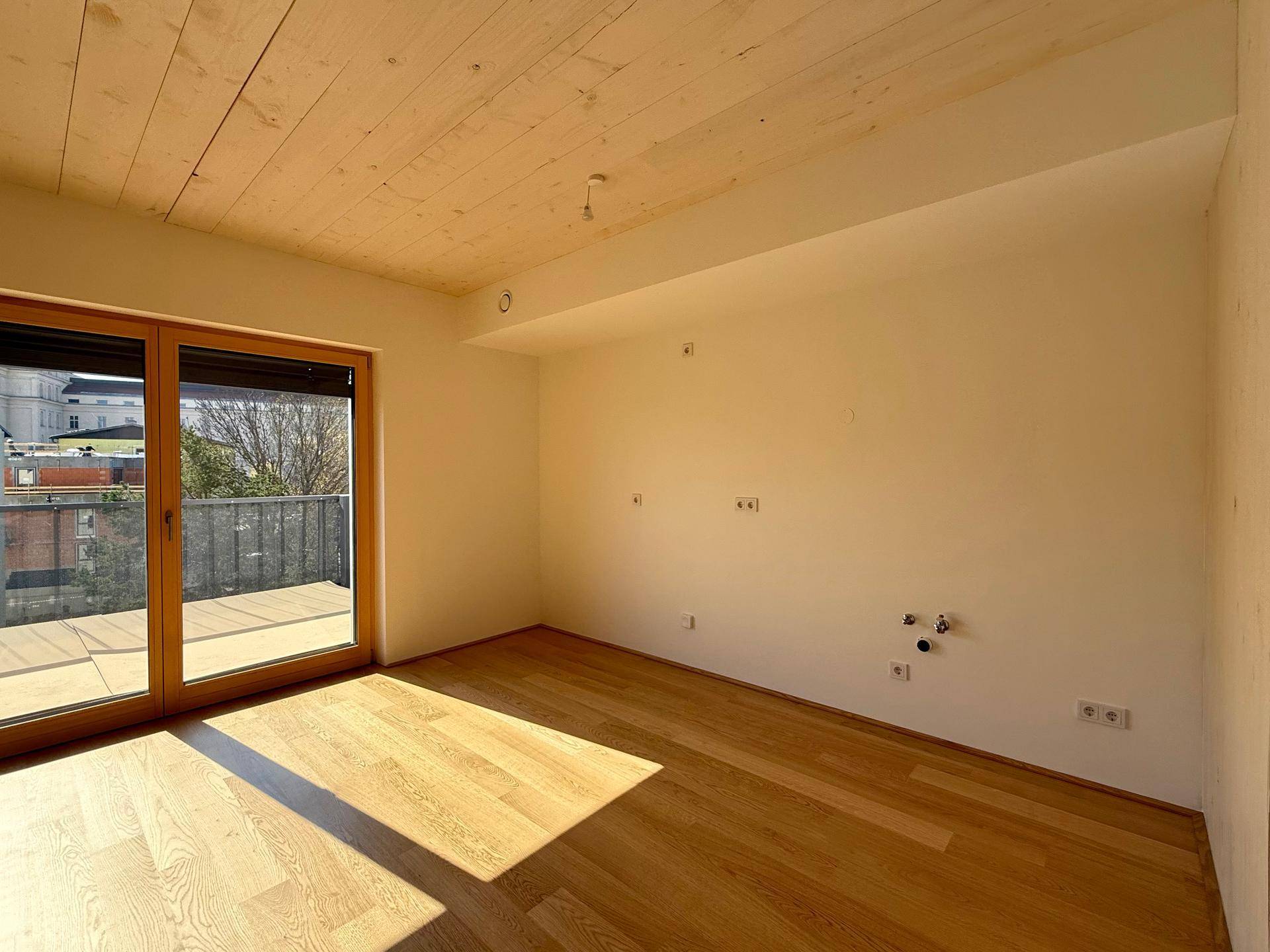Helles Zimmer mit Holzboden und Balkonzugang, vorbereitet für eine mögliche Kücheninstallation.