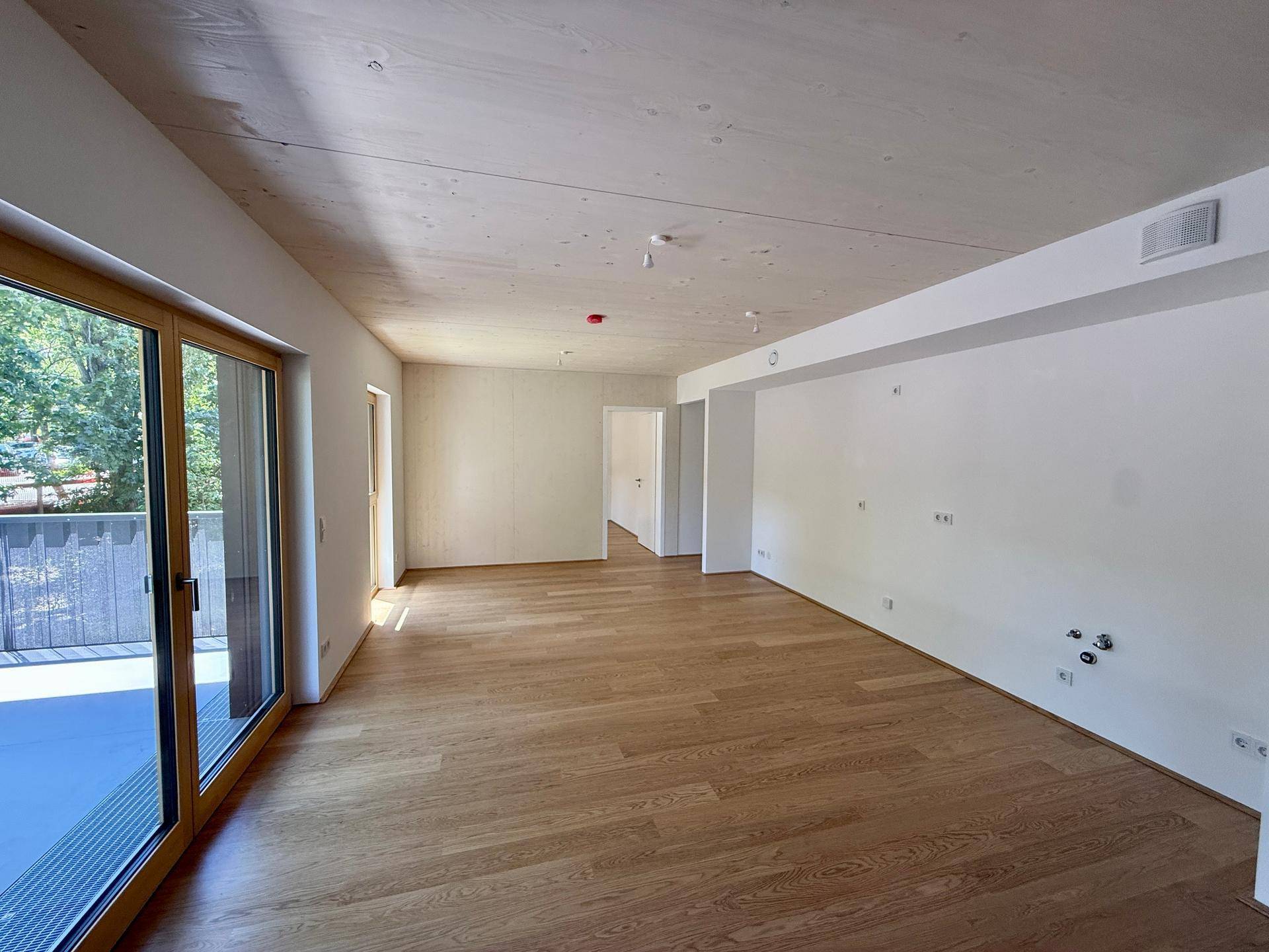 Leerer, lichtdurchfluteter Raum mit Holzboden, hohen Decken und großen Fenstern zum Balkon.