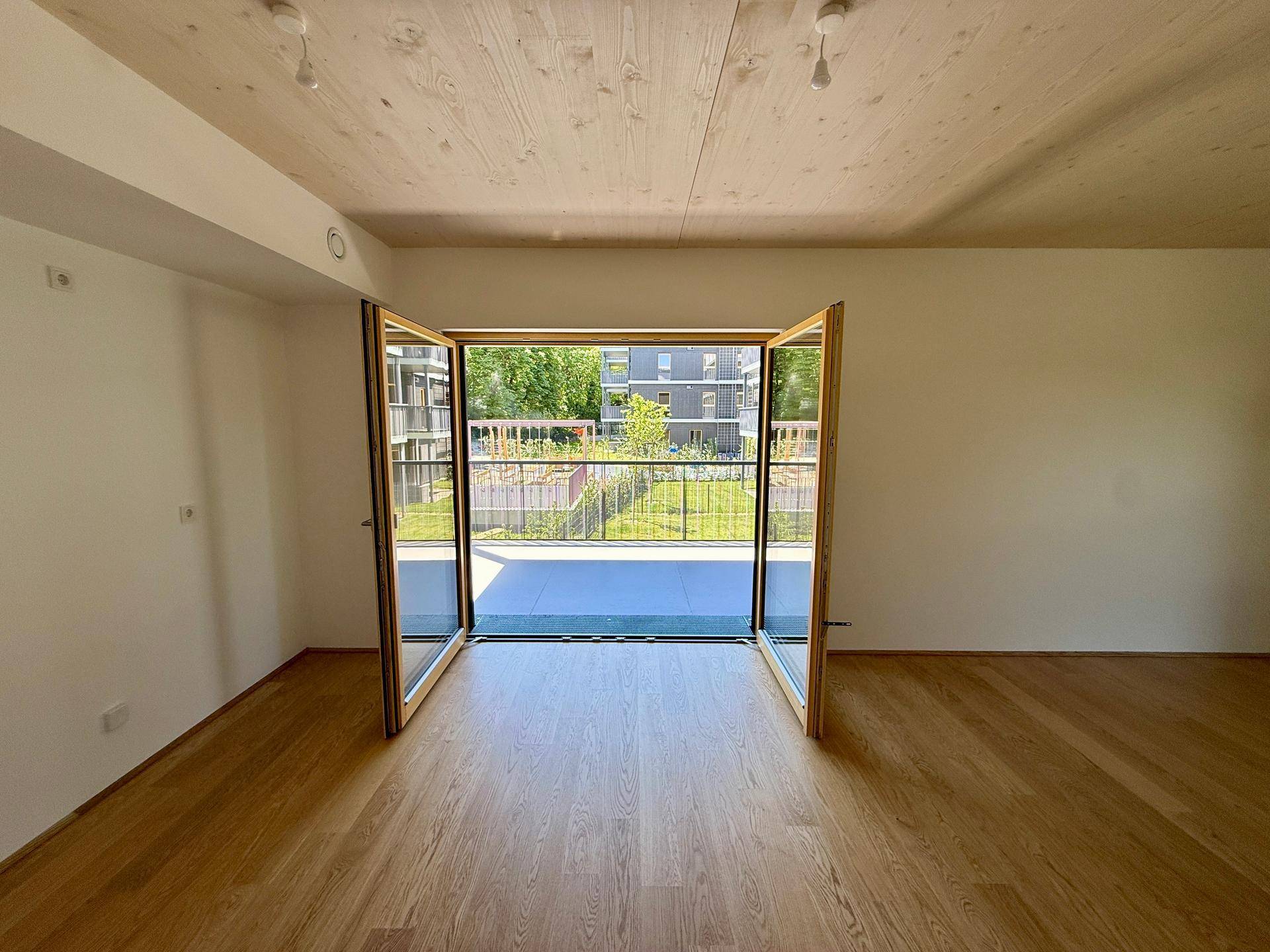 Lichtdurchfluteter Wohnbereich mit Holzboden und Zugang zu einem Balkon mit Blick auf die Außenanlage.