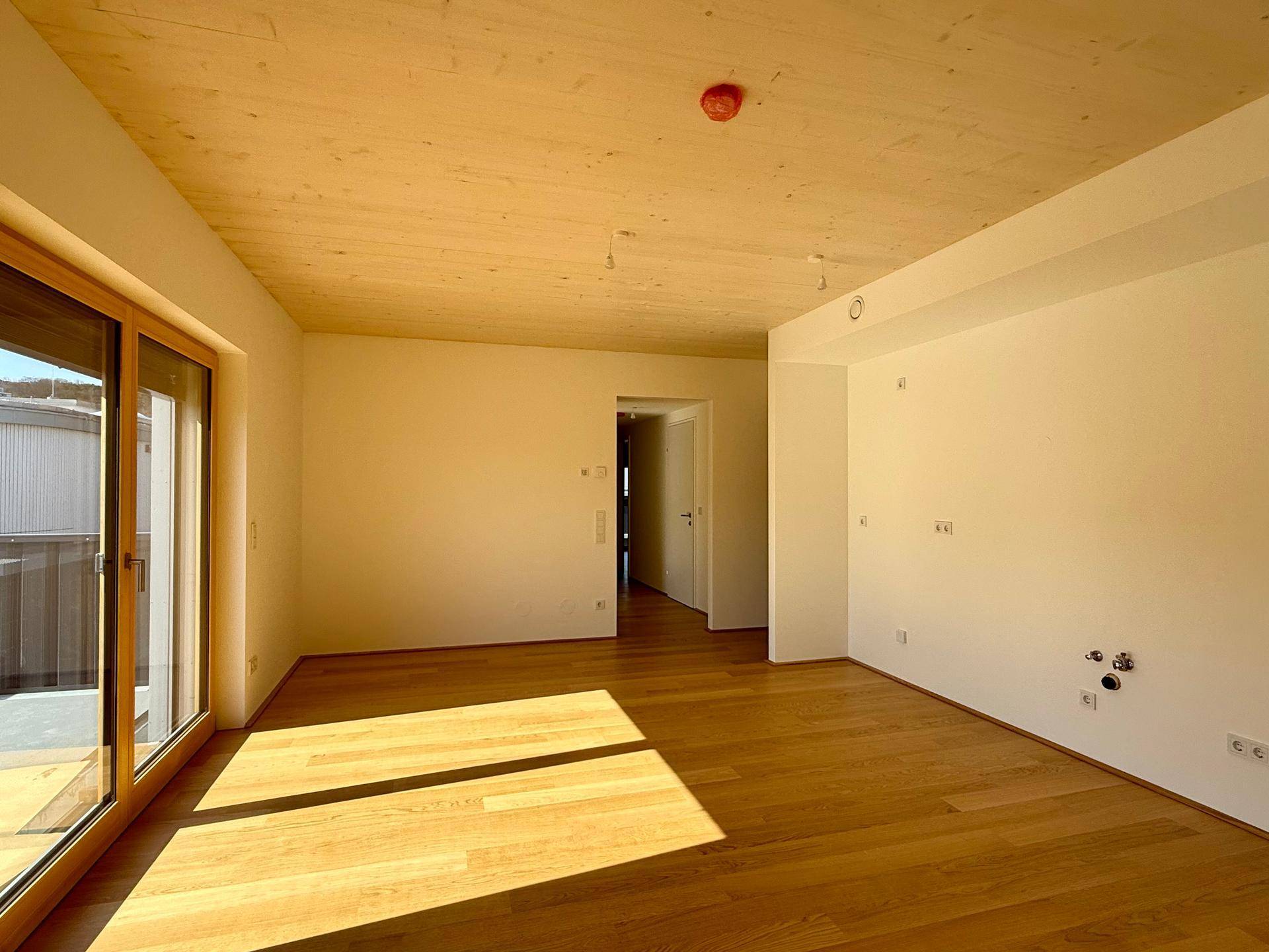 Heller Wohnbereich mit Holzboden und vorbereiteten Anschlüssen für eine mögliche Küchenzeile.
