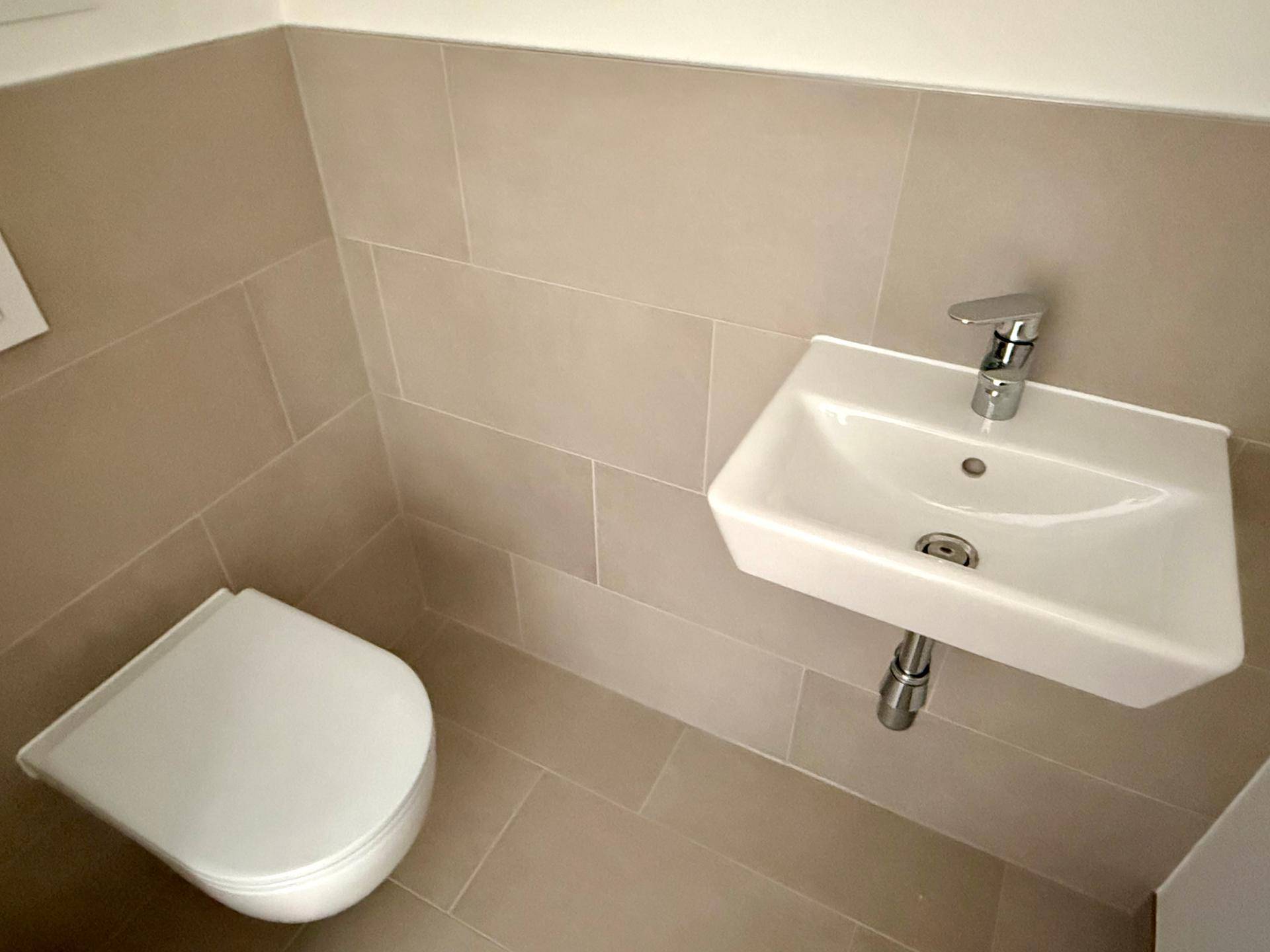 Modernes Gäste-WC mit beigefarbenen Fliesen, wandhängendem WC und kleinem Waschbecken.