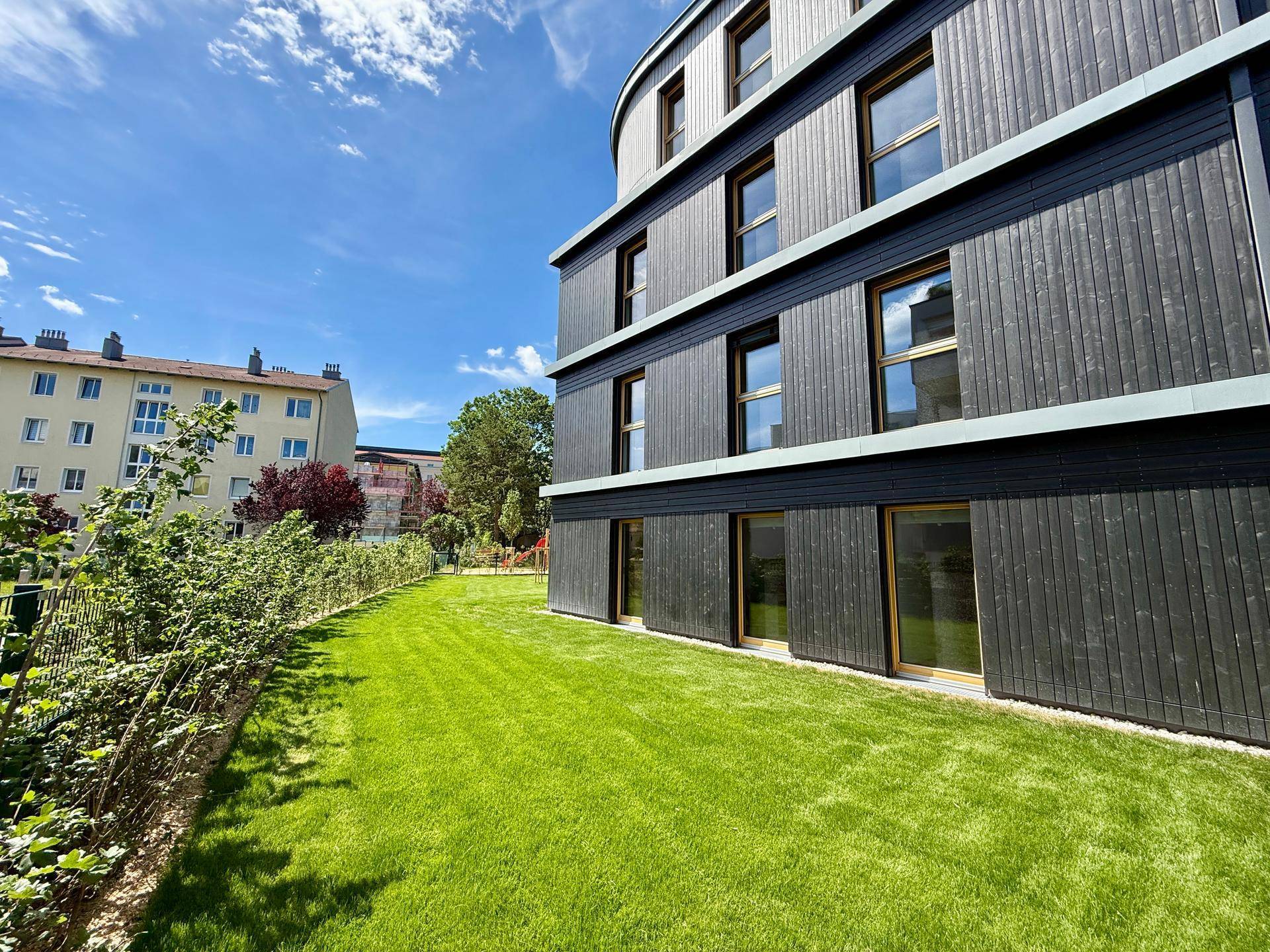 Ansicht der modernen Wohnanlage mit dunkler Holzfassade und weitläufiger Grünfläche unter blauem Himmel.