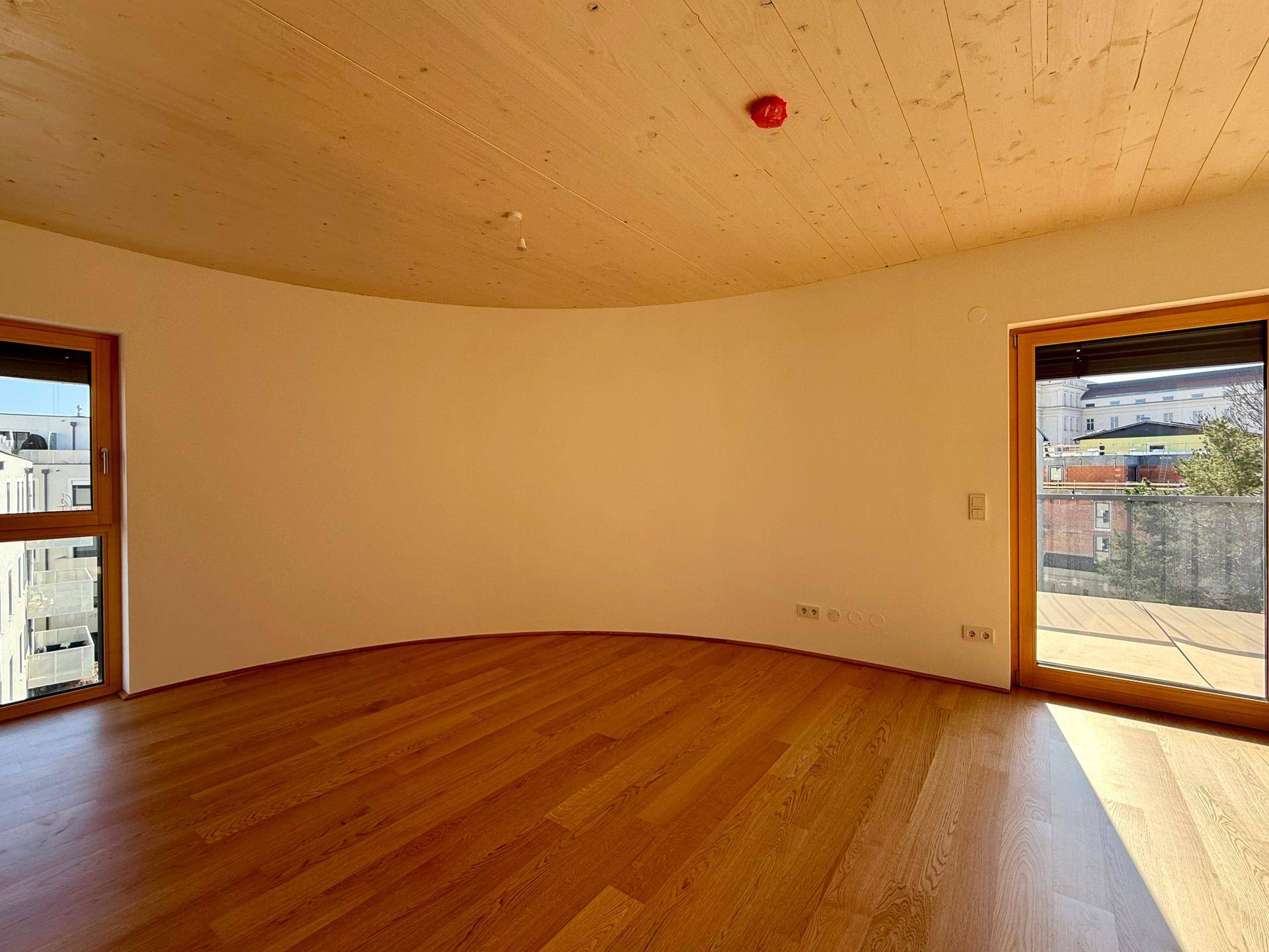 Heller Raum mit geschwungener Wand, Holzboden und Zugang zu einem Außenbereich.