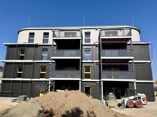 Neubau-Fassade mit dunklen Paneelen und mehreren Balkonen, umgeben von Baustellengelände.