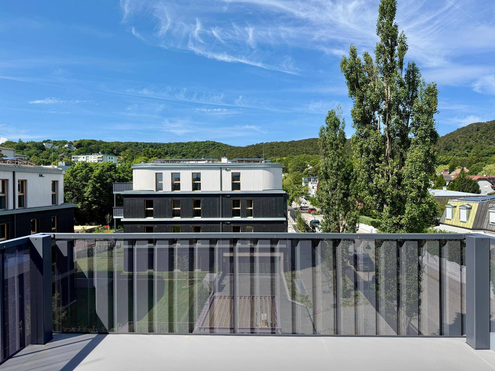 Balkon mit Blick auf ein modernes Gebäude, hohe Bäume und die bewaldeten Hügel in der Ferne.