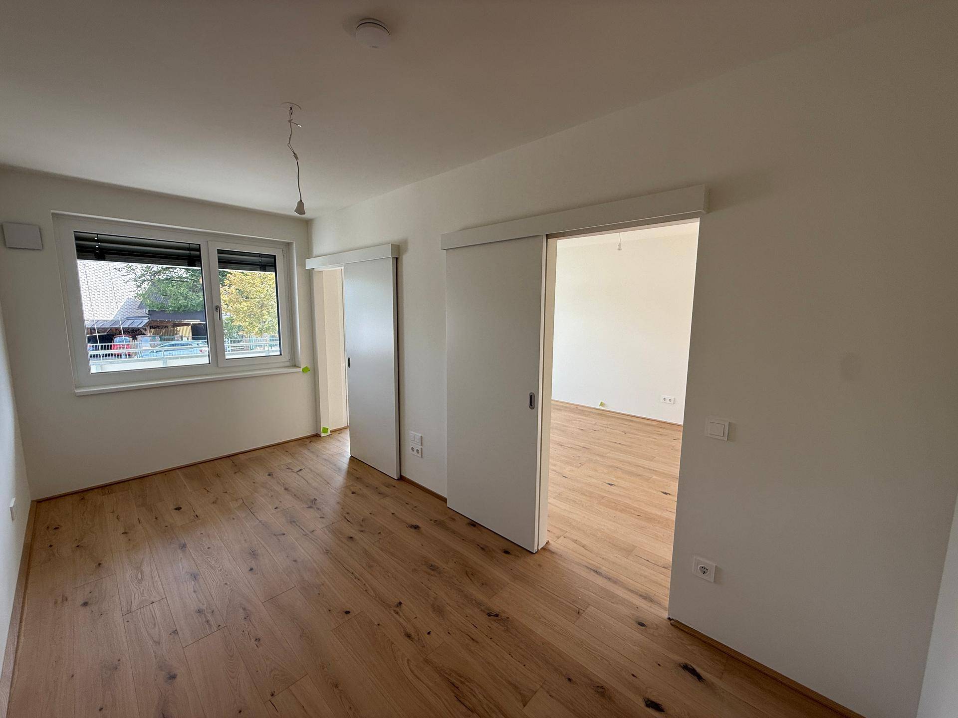 Unmöbliertes Zimmer mit Holzboden, Fenster und zwei modernen Schiebetüren.
