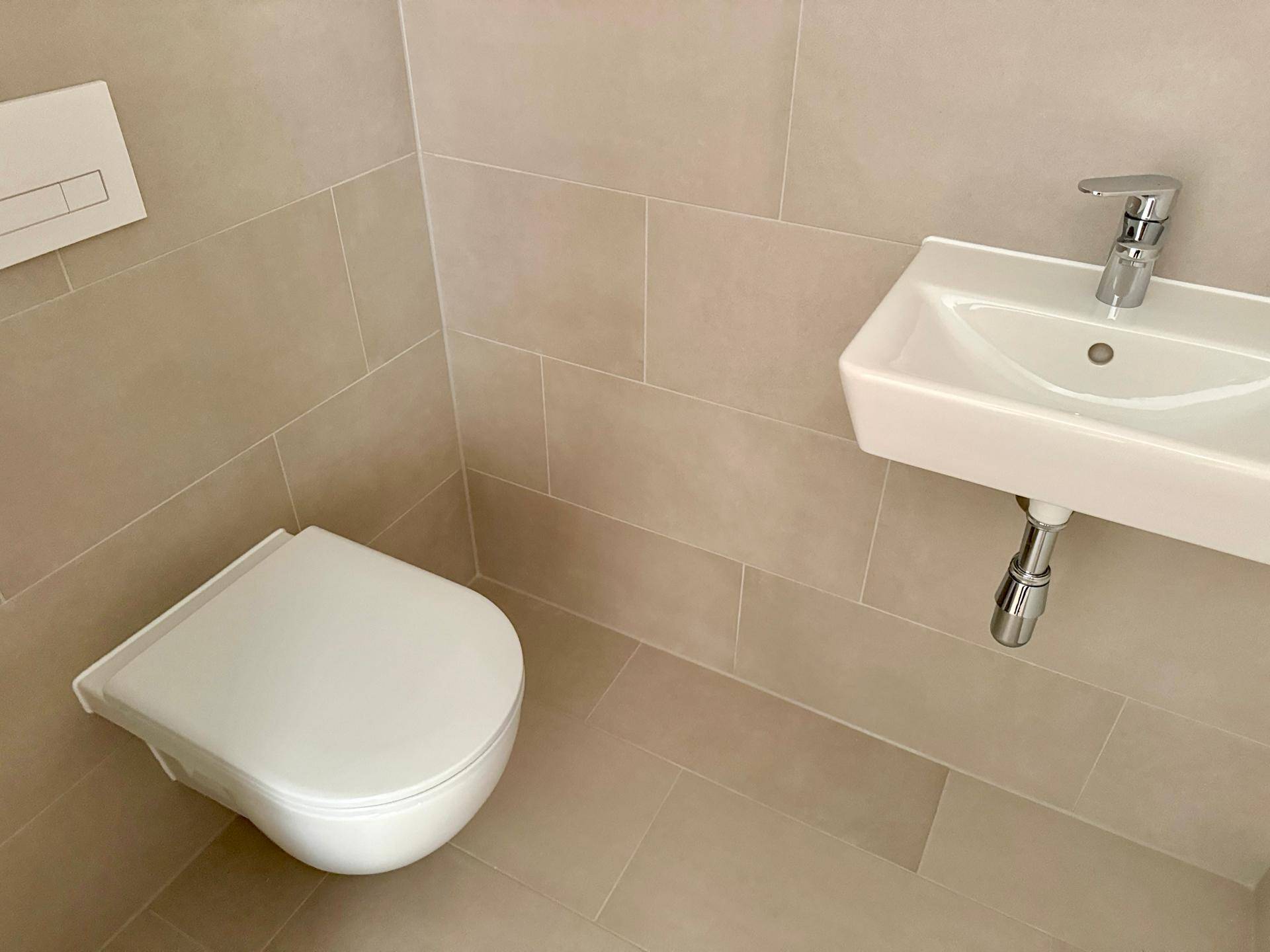 Gäste-WC mit wandhängender Toilette, kleinem Waschbecken und beigefarbenen Fliesen.