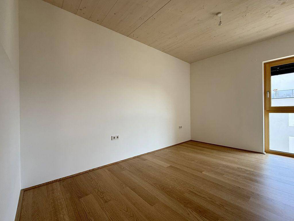 Helles Zimmer mit Holzboden und großem Fenster, ideal als Schlaf- oder Arbeitszimmer in einem Neubau.