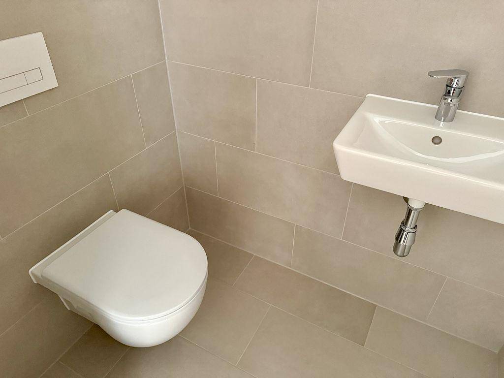 Separate Toilette mit Wand-WC, kleinem Waschbecken und beigefarbenen Wandfliesen.