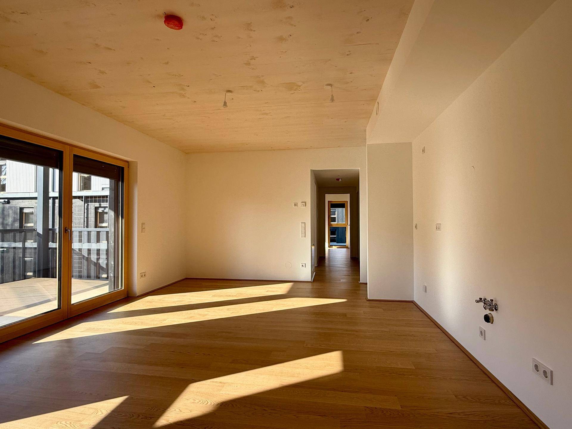 Geräumiges Wohnzimmer mit Holzboden und hellem Anstrich, Blick in den Flur und Zugang zum Außenbereich.