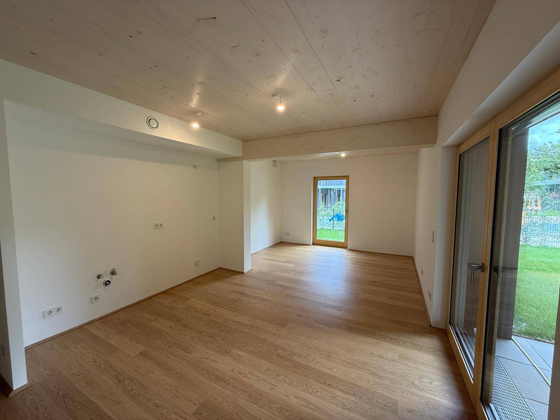 Heller, leerer Raum mit Holzboden und Holzdecke, großen Fenstern und Zugang zum Garten.