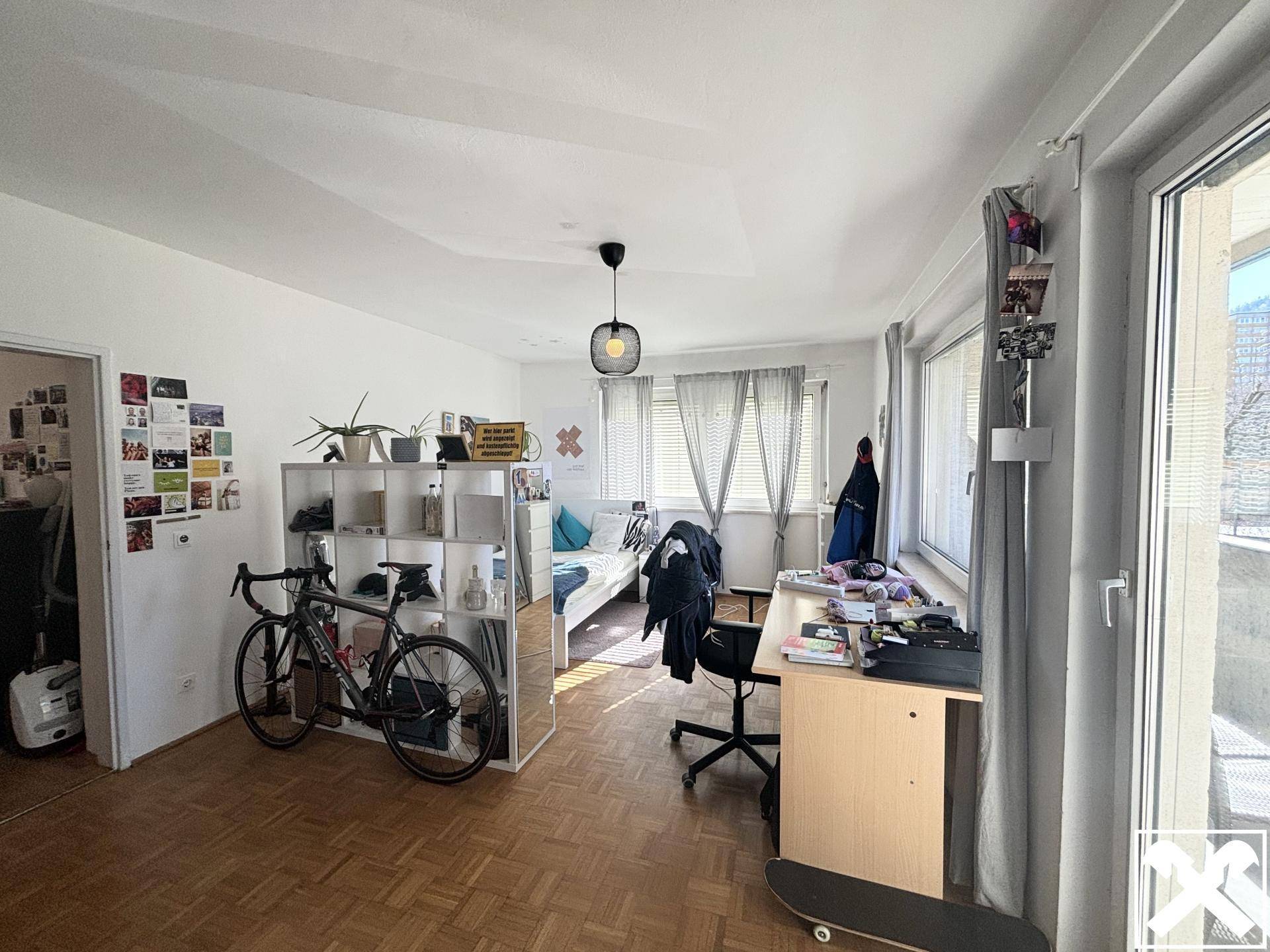 Helles Zimmer mit Parkettboden, Schreibtisch und Zugang zu einem kleinen Balkon.