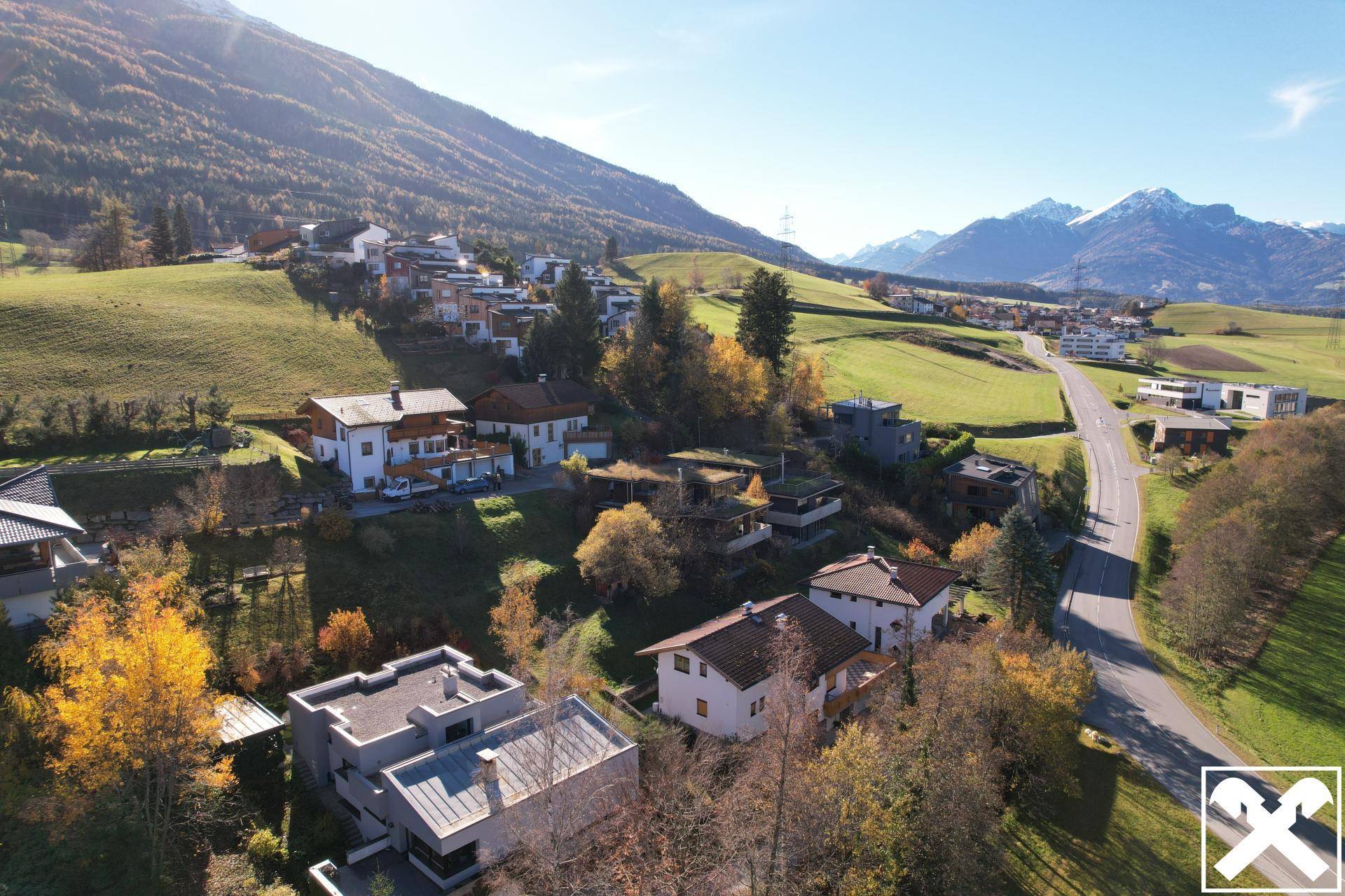 Weitläufiger Blick auf ein Wohngebiet mit Straße, Feldern und einer beeindruckenden Bergkulisse.