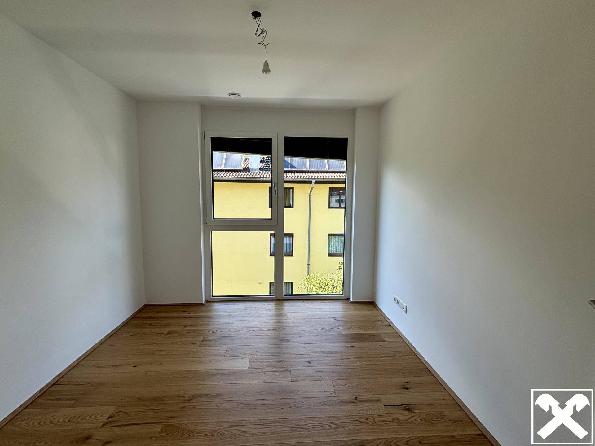 Unmöbliertes Schlafzimmer mit hellem Holzparkett und großem Fenster mit Blick auf Nachbargebäude.