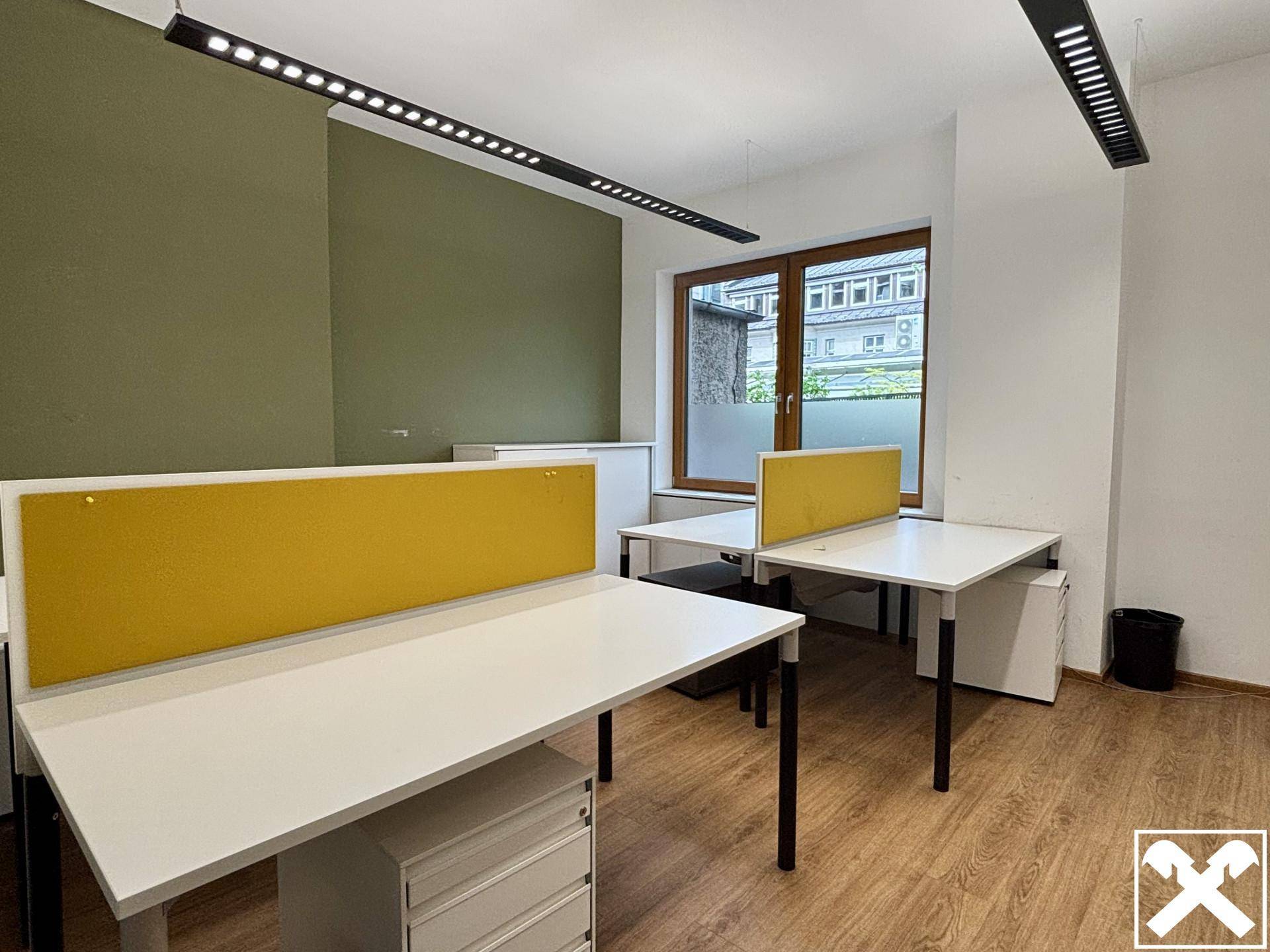 Offener Bürobereich mit mehreren Schreibtischen, gelben Trennwänden und einer grünen Akzentwand, hell und modern.