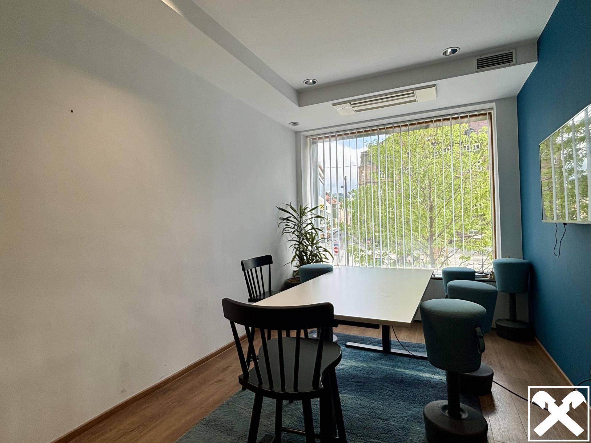 Besprechungsbereich mit rechteckigem Tisch, schwarzen Stühlen und blauer Akzentwand, gut beleuchtet durch ein Fenster.